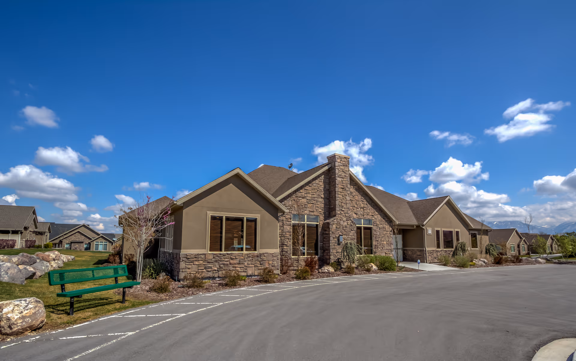 Exterior view of a single-story building with stone and beige siding under a bright blue sky with scattered clouds. There is a green bench on the grass near the building and a paved driveway in front.
