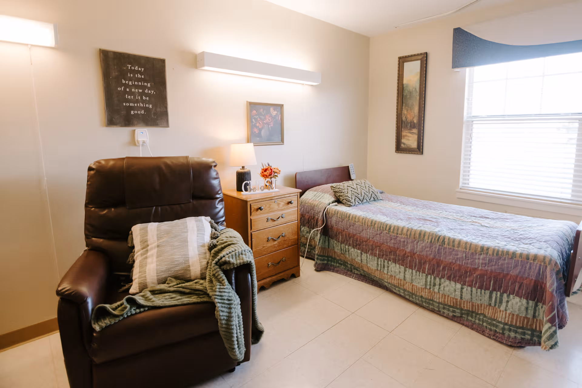 A furnished bedroom with a single bed, recliner chair, dresser, and soft lighting.