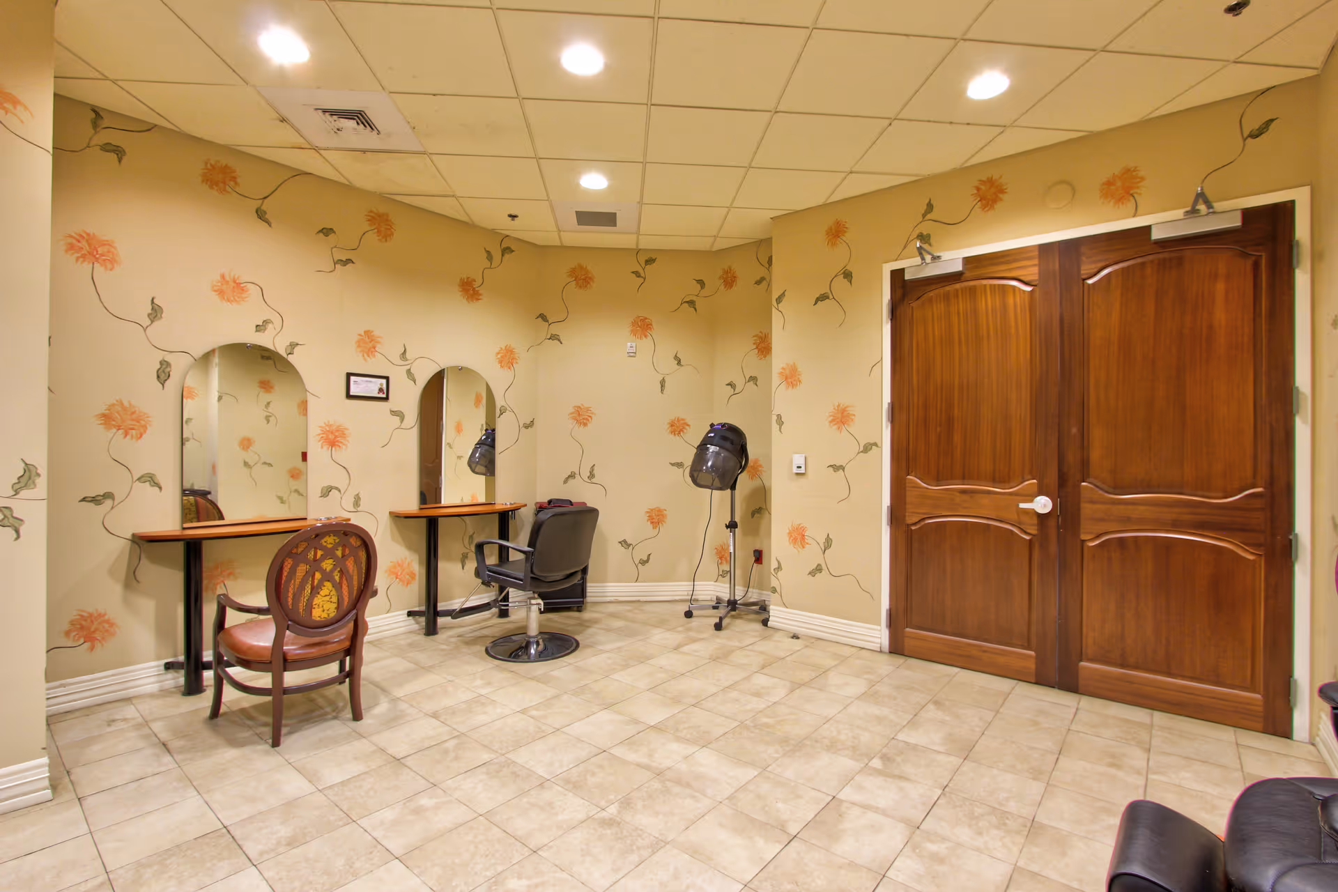 Interior view of a hair salon area in a senior living facility with two styling stations, each with a mirror and chair. One chair is a wooden armchair with a patterned backrest, and the other is a black salon chair. A hair dryer on a stand is positioned near the corner. The walls are decorated with floral wallpaper, and there are double wooden doors on the right side.