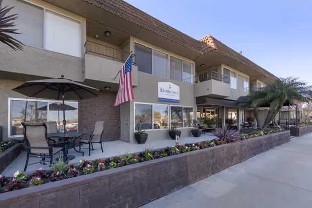 Exterior view of Brookdale Valley View facility showing a two-story building with balconies, an American flag, outdoor seating with umbrellas, and a flower bed along the sidewalk.