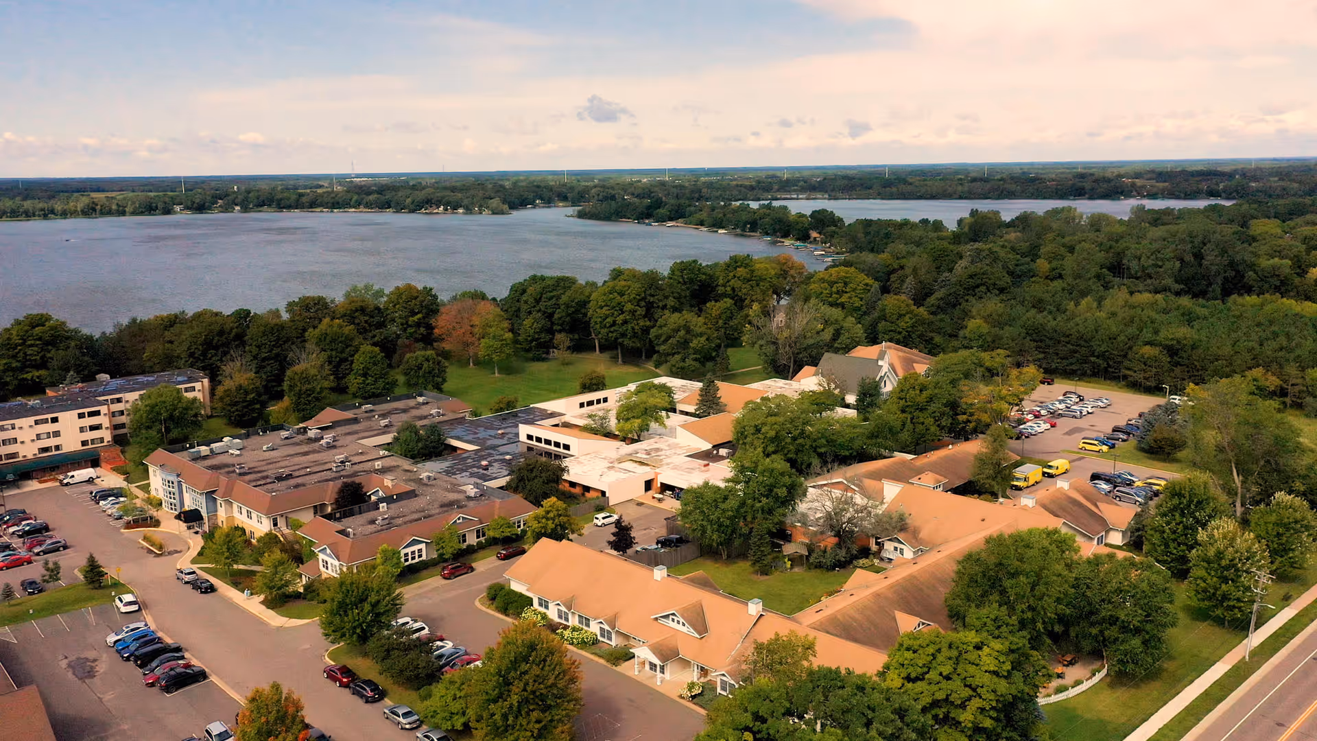 Aerial view of Parmly on the Lake senior living facility surrounded by trees and greenery, with a large lake in the background and several parking areas filled with cars.