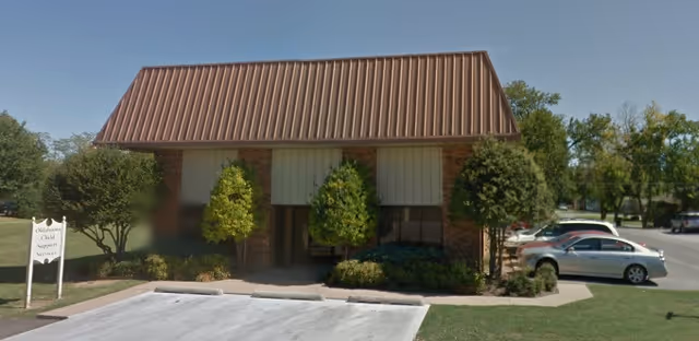 Front view of a brick medical facility with brown metal roof, shrubs, and a parked car.