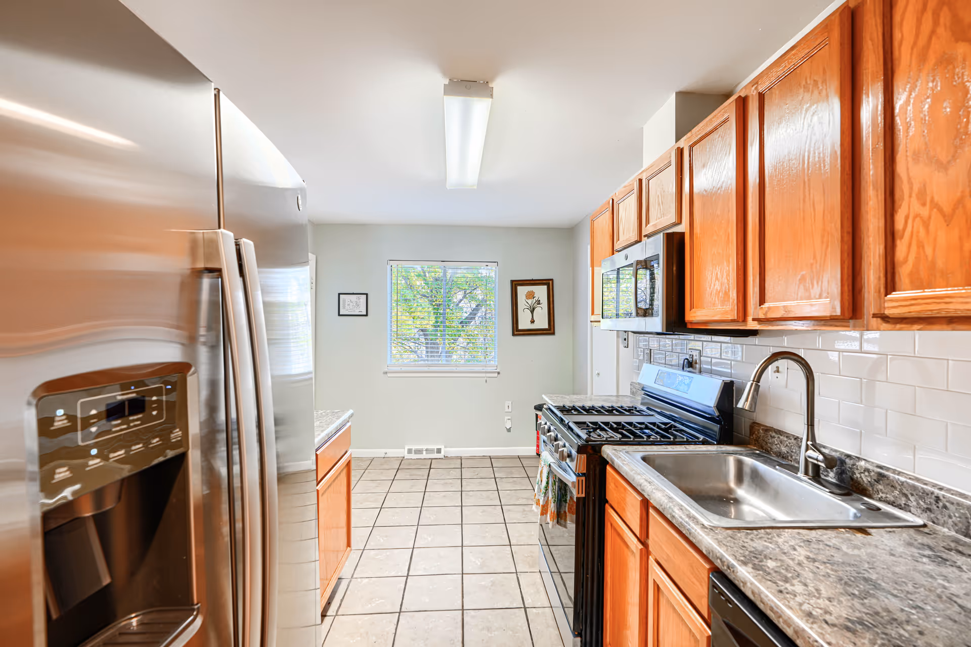 Bright kitchen with stainless fridge, wooden cabinets, stove, sink, and tiled floor.