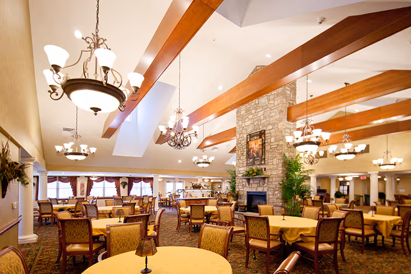 Spacious dining room with multiple round tables covered with yellow tablecloths and surrounded by wooden chairs with patterned upholstery. The room features a high ceiling with wooden beams and several ornate chandeliers. A large stone fireplace with plants on either side is visible in the center background. Large windows with curtains allow natural light to fill the room.