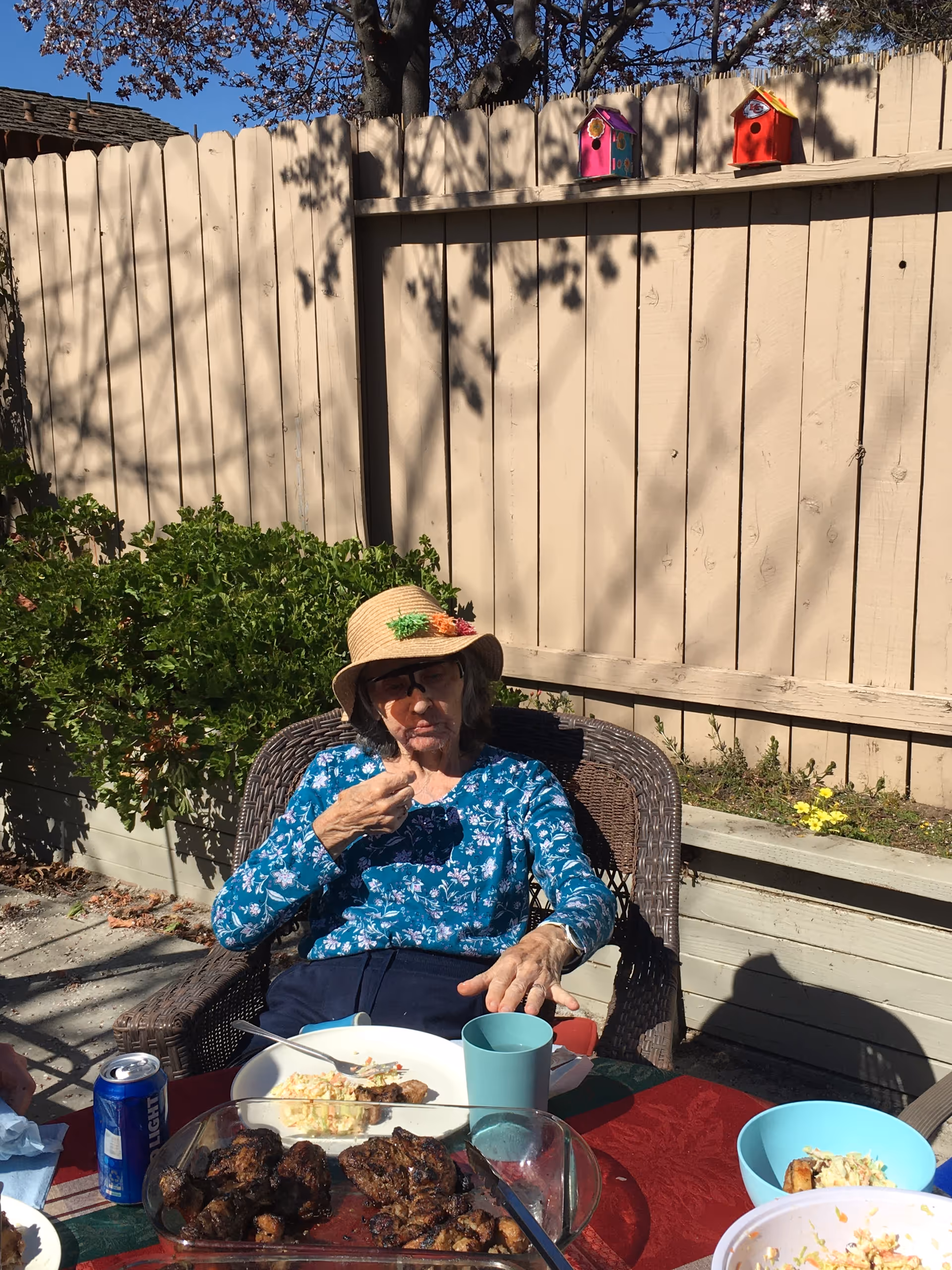 Elderly woman wearing a sun hat sits at an outdoor table with food and drinks.