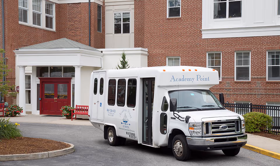 A white shuttle bus labeled 'Academy Point' parked in front of a brick building with red double doors and white columns at the entrance. There are red benches and some greenery around the entrance area.