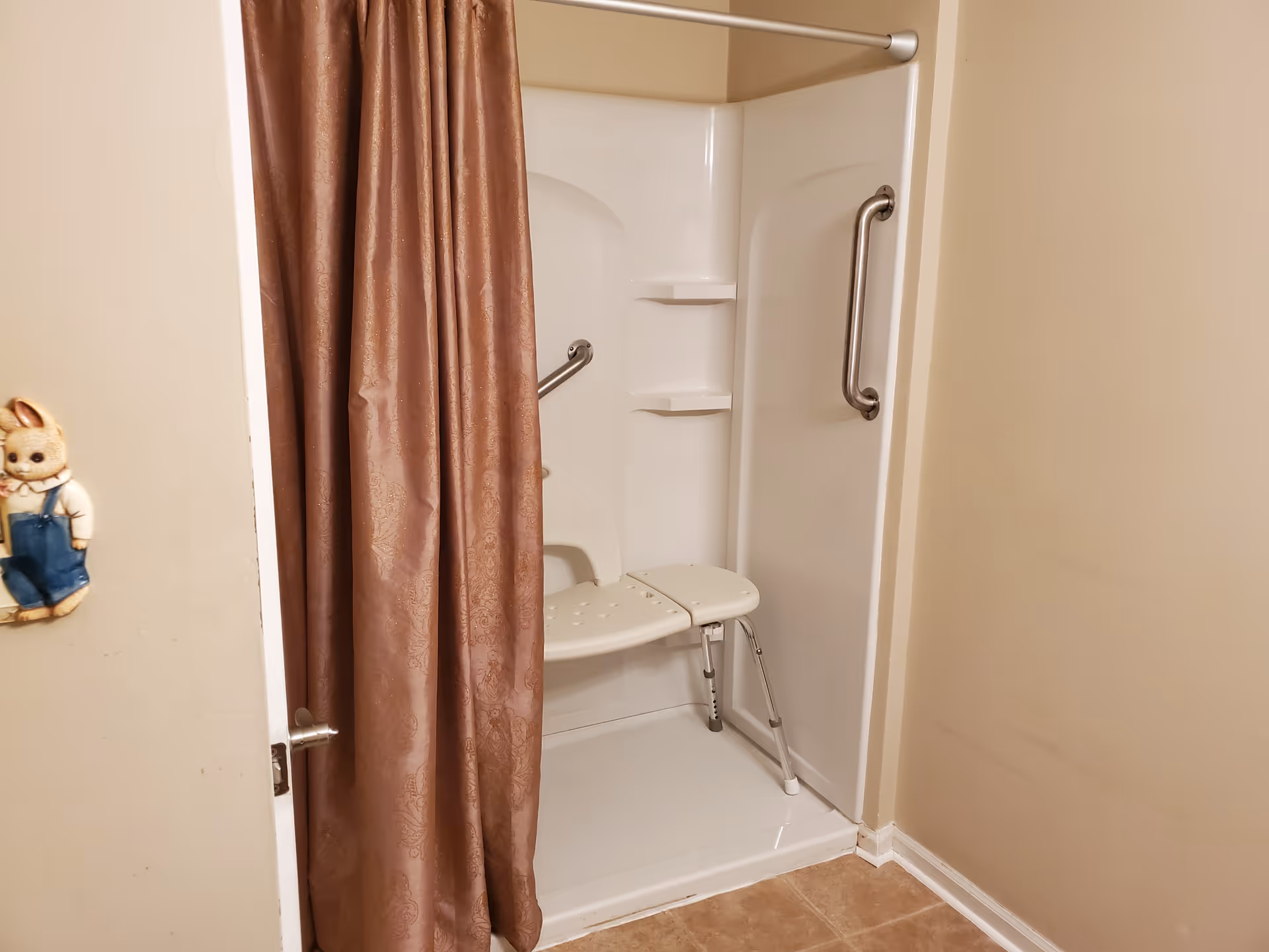 Fold-down shower seat with grab bars and brown curtain in a beige bathroom.