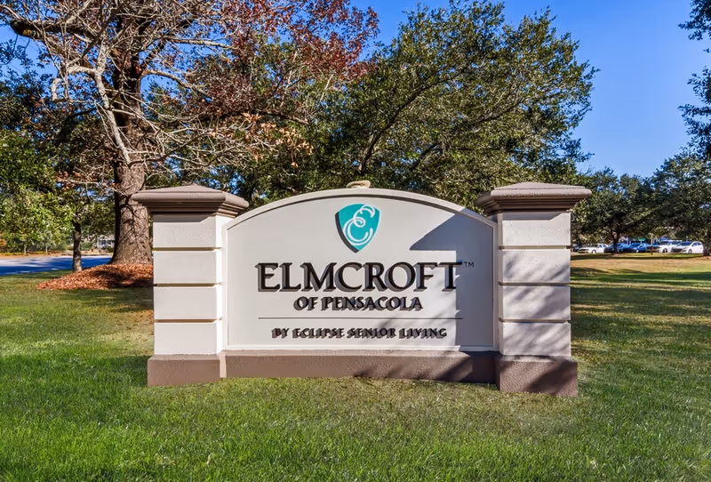 Outdoor view of a large stone sign for Elmcroft of Pensacola by Eclipse Senior Living, surrounded by green grass and trees under a clear blue sky.