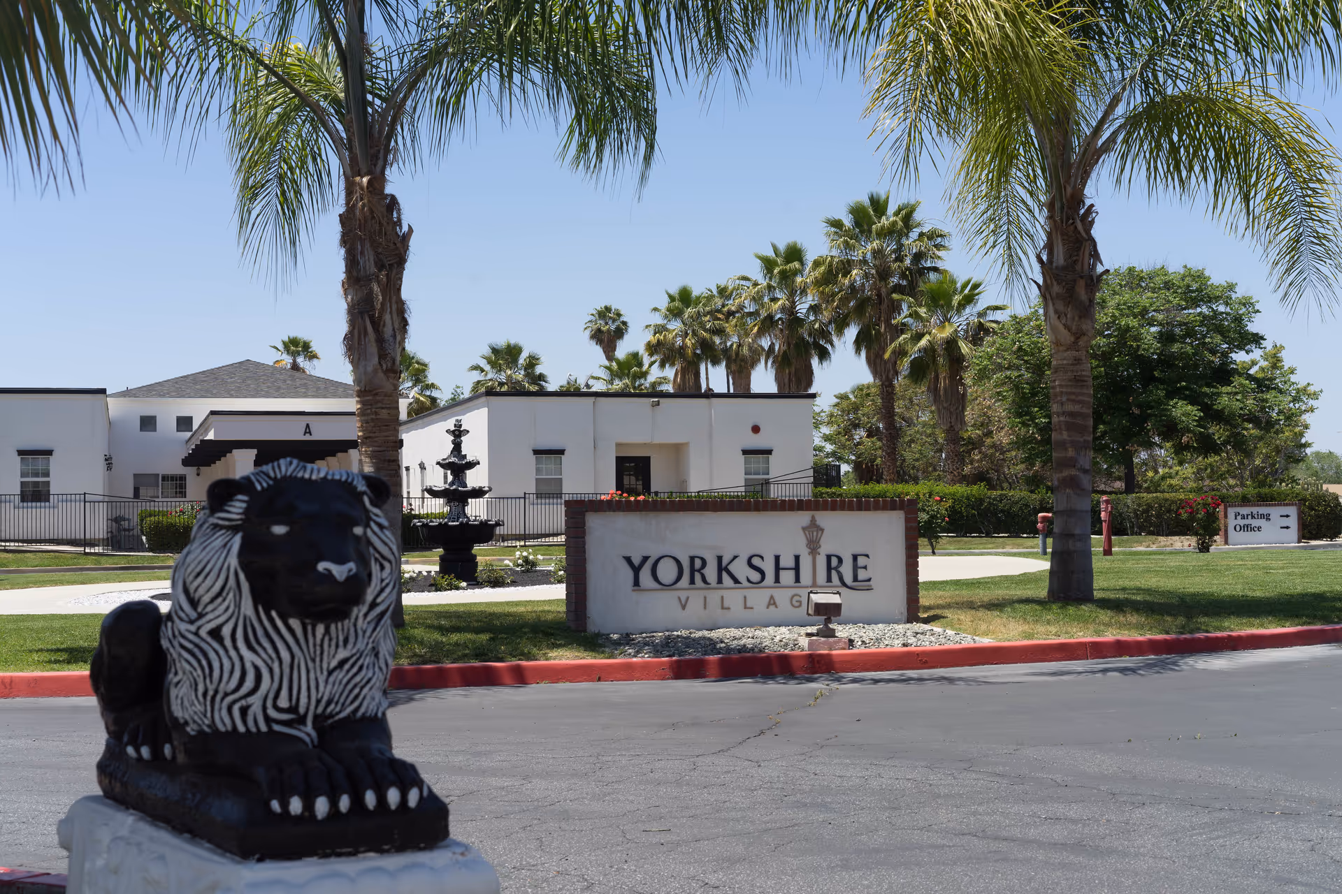 Exterior view of Yorkshire Village facility with a sign, palm trees, statue, and low fountain