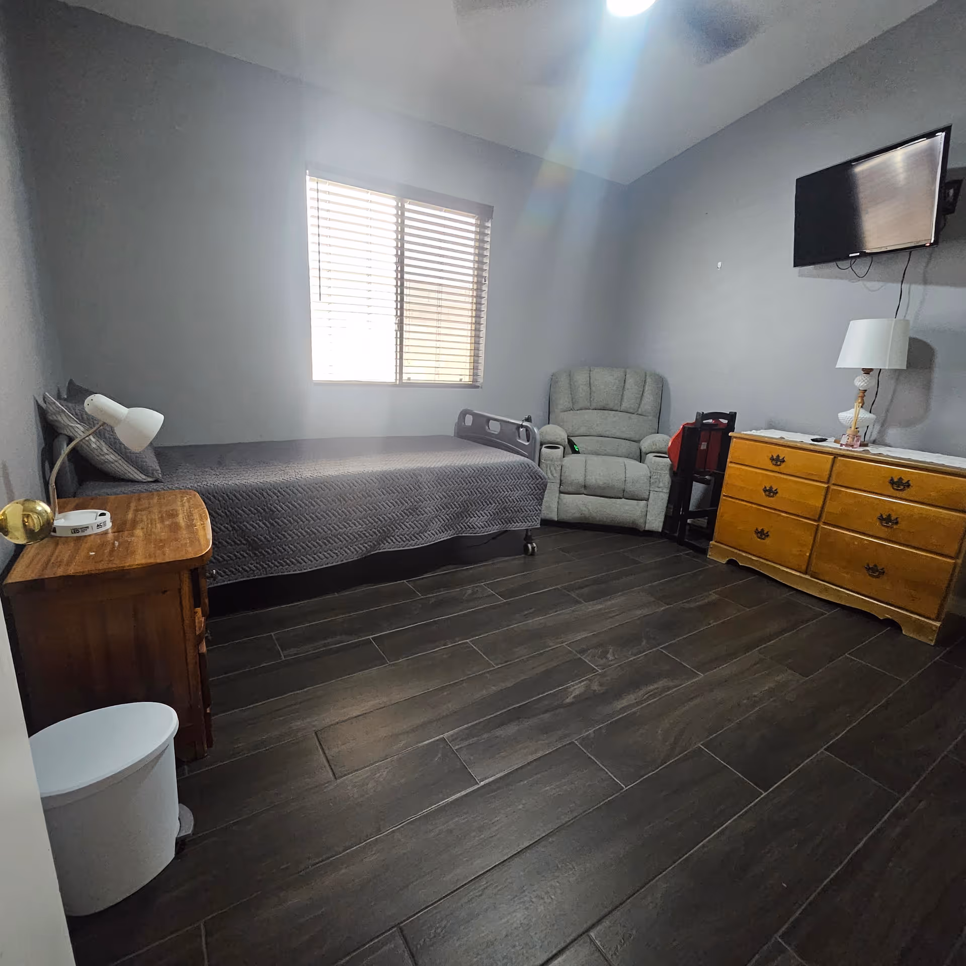 A modest bedroom in a senior facility with a single bed, dresser, chair, and TV.
