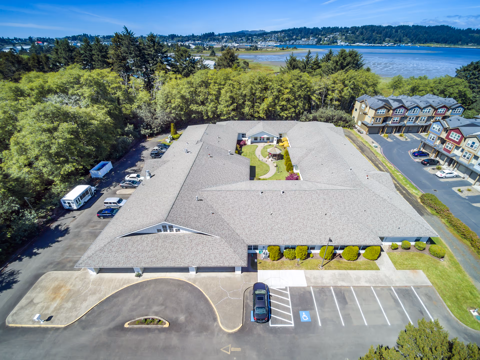 Aerial view of South Beach Manor Memory Care facility showing a large single-story building with a central courtyard featuring a walking path and gazebo. The building is surrounded by parking spaces, some vehicles, and greenery with trees and a body of water in the background.