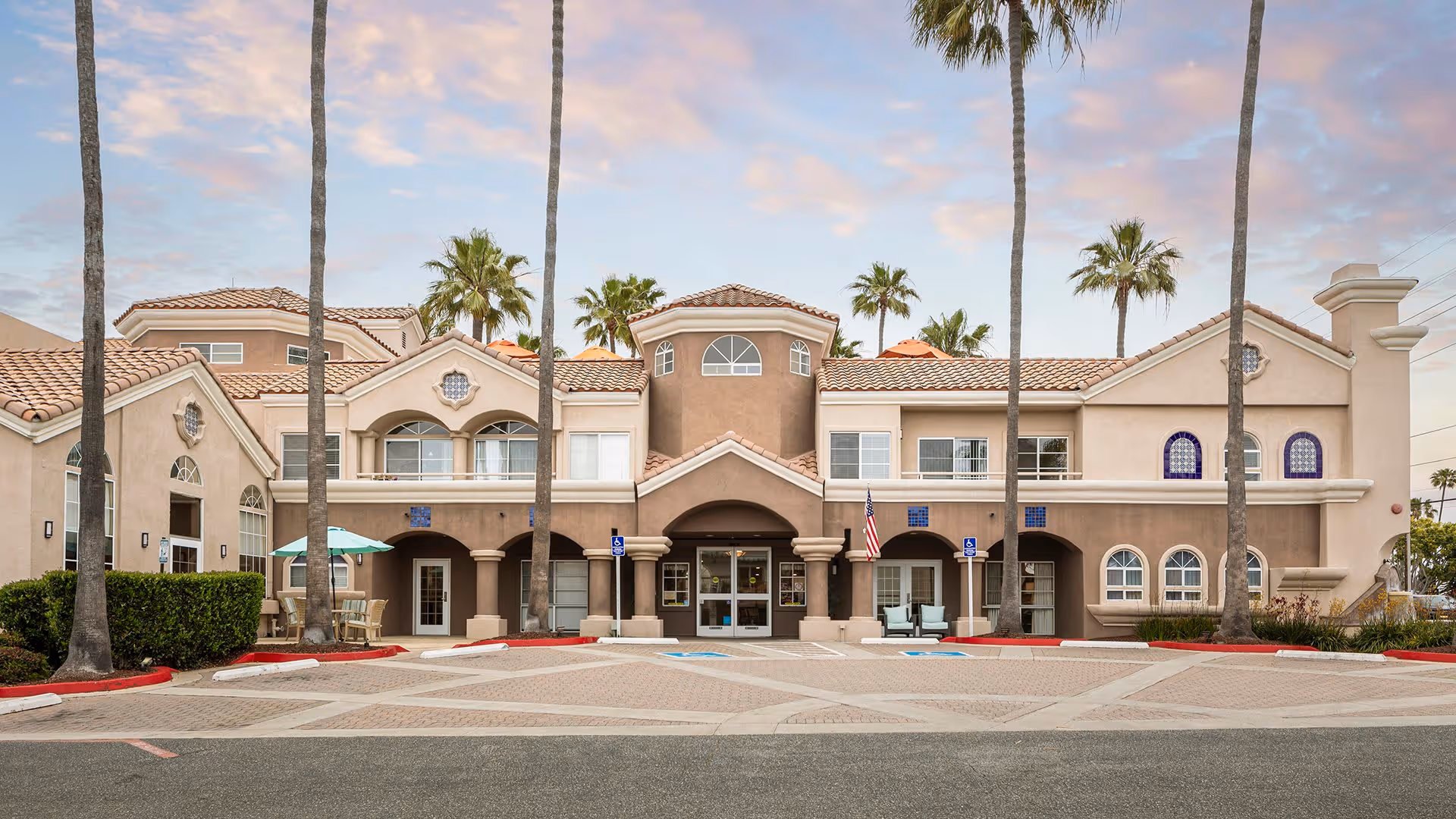 Front exterior view of Atria Newport Plaza building with palm trees, a paved parking area with handicapped spaces, and a partly cloudy sky at sunset.