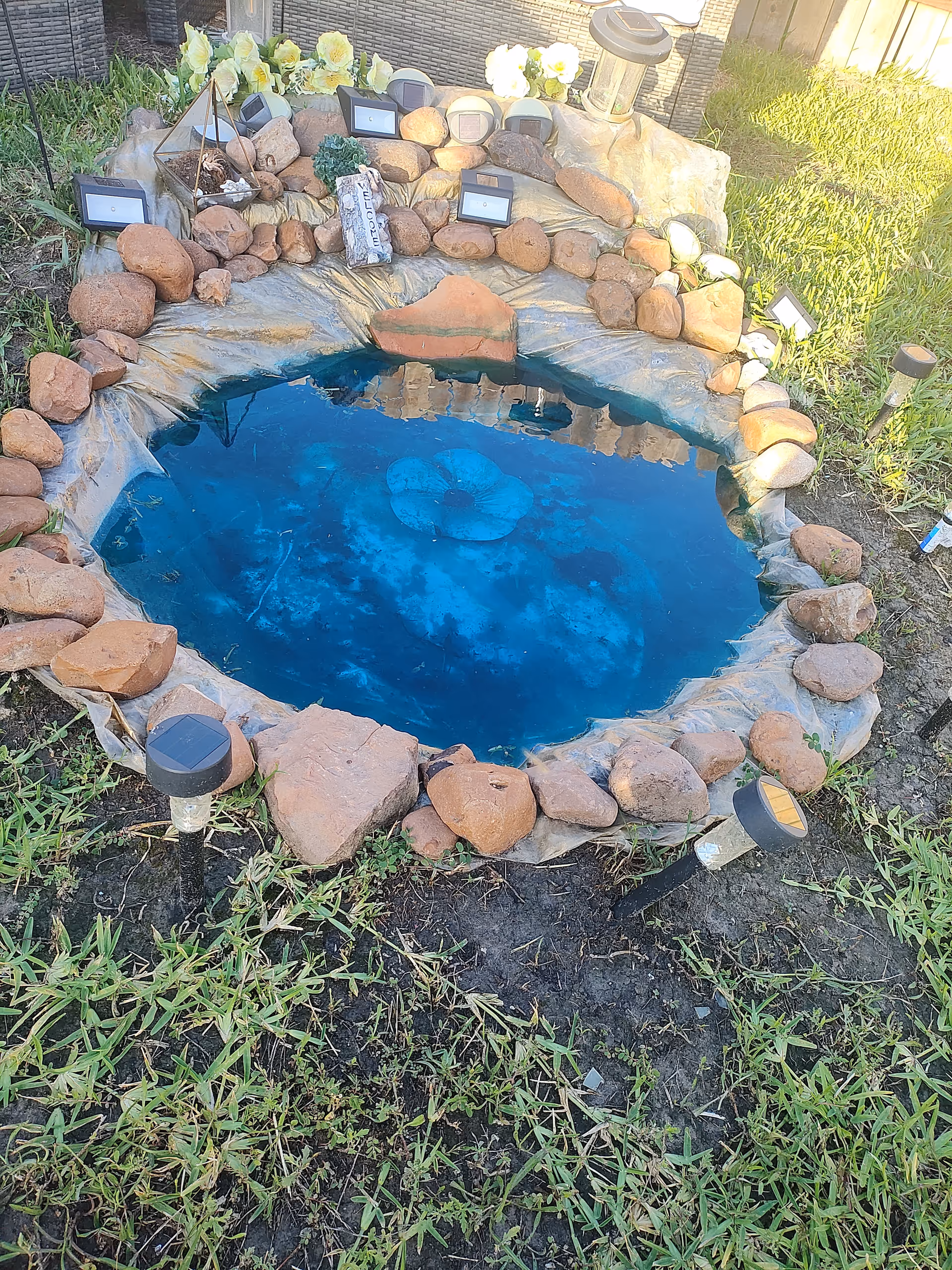 Circular stone-bordered backyard pond with blue water, rocks, and landscape lights.