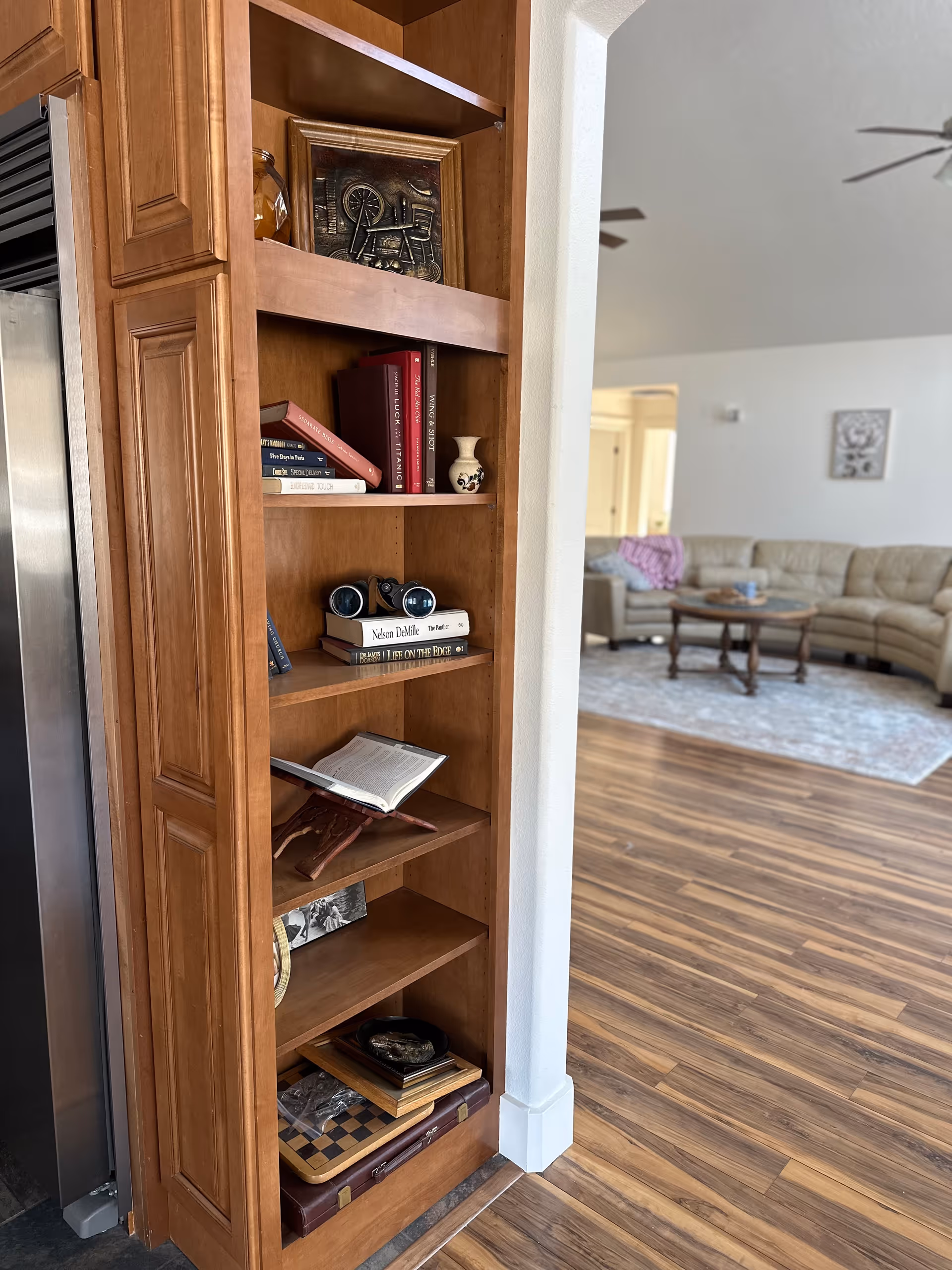 Tall wooden bookshelf with books and decor beside the kitchen, opening to a living room.