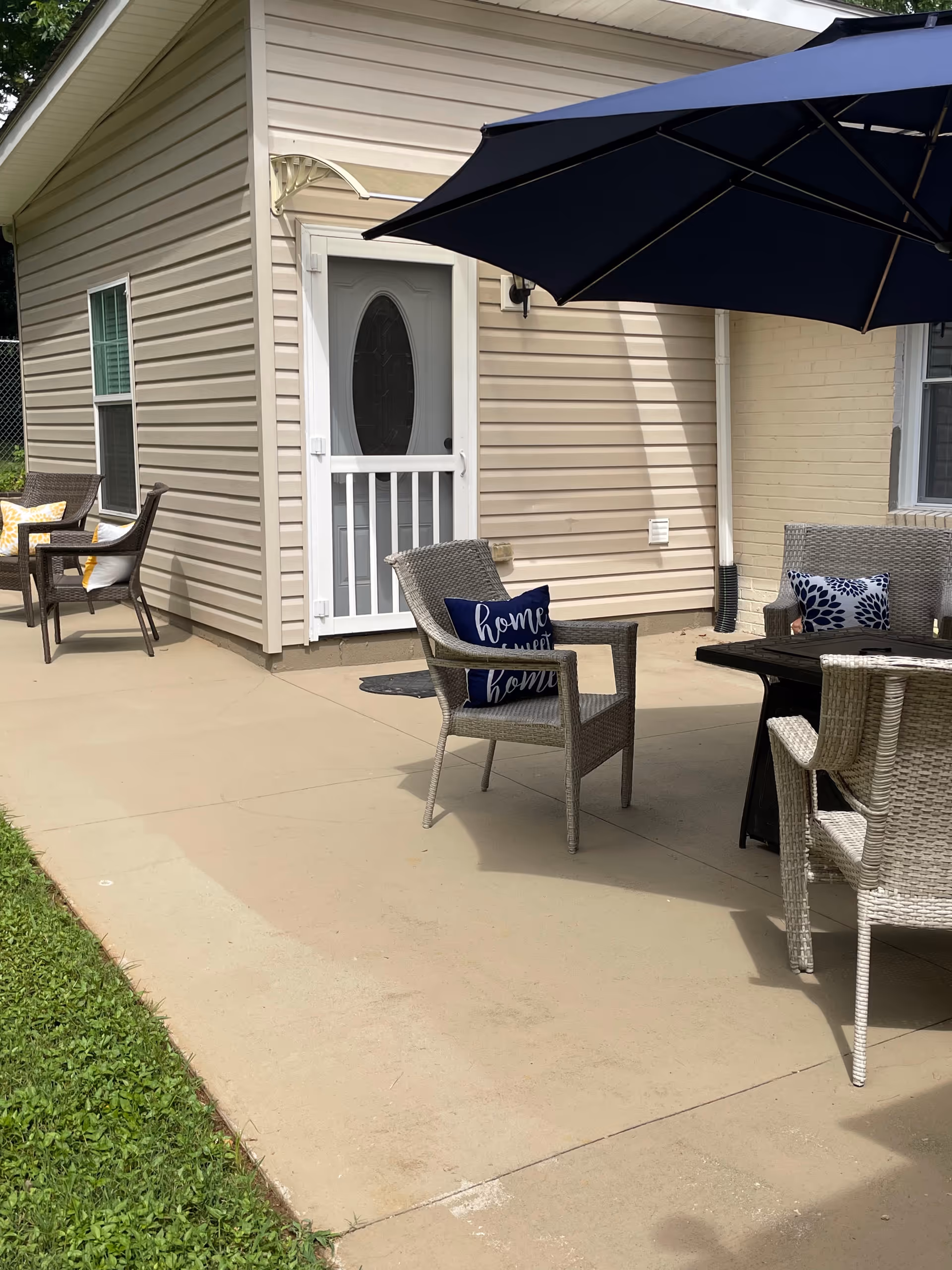 Outdoor patio with wicker chairs, a table, cushions, and a large umbrella beside a beige building.