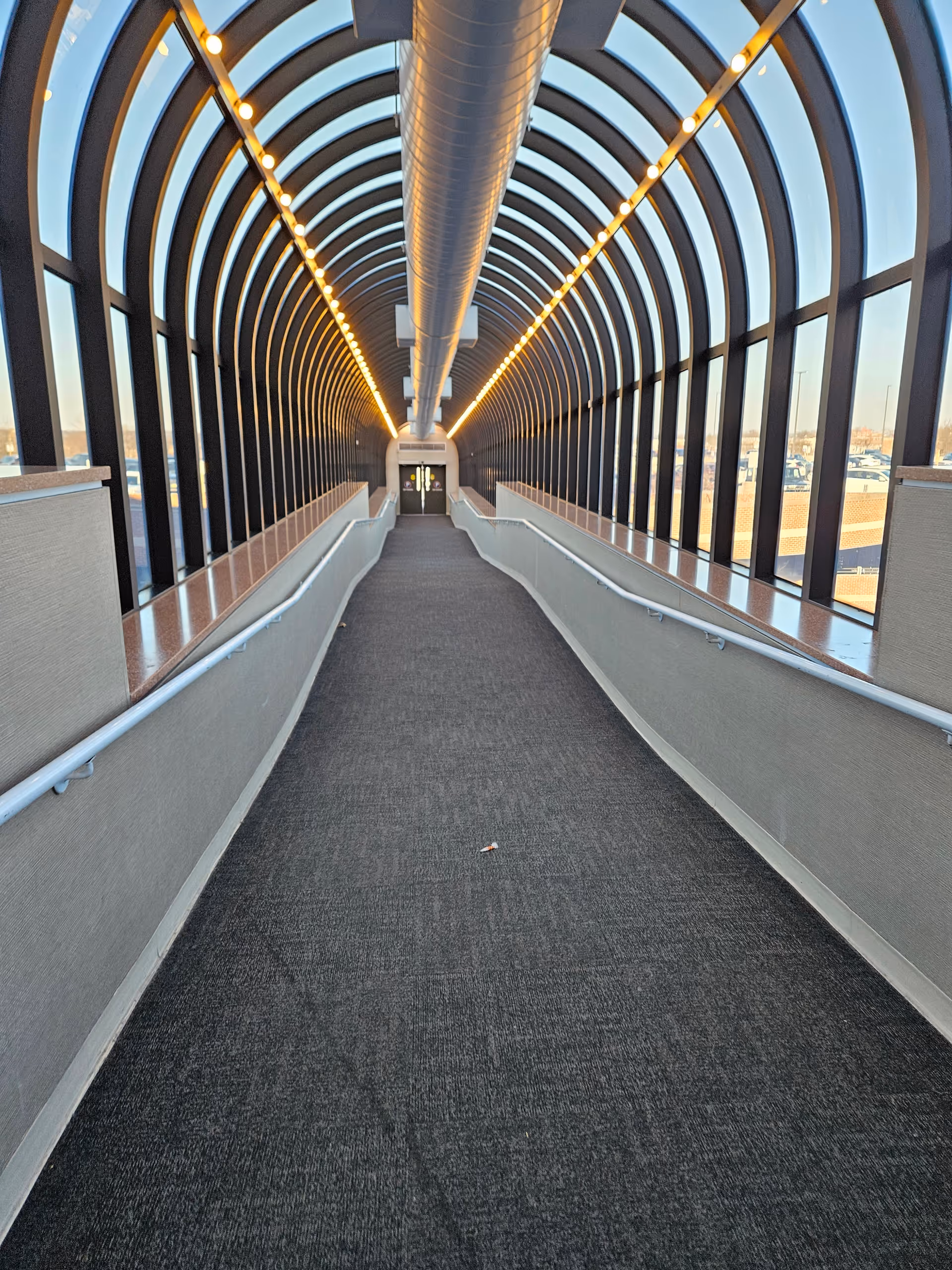 Glass-enclosed indoor walkway with arched framing, warm lighting, and carpeted floor.