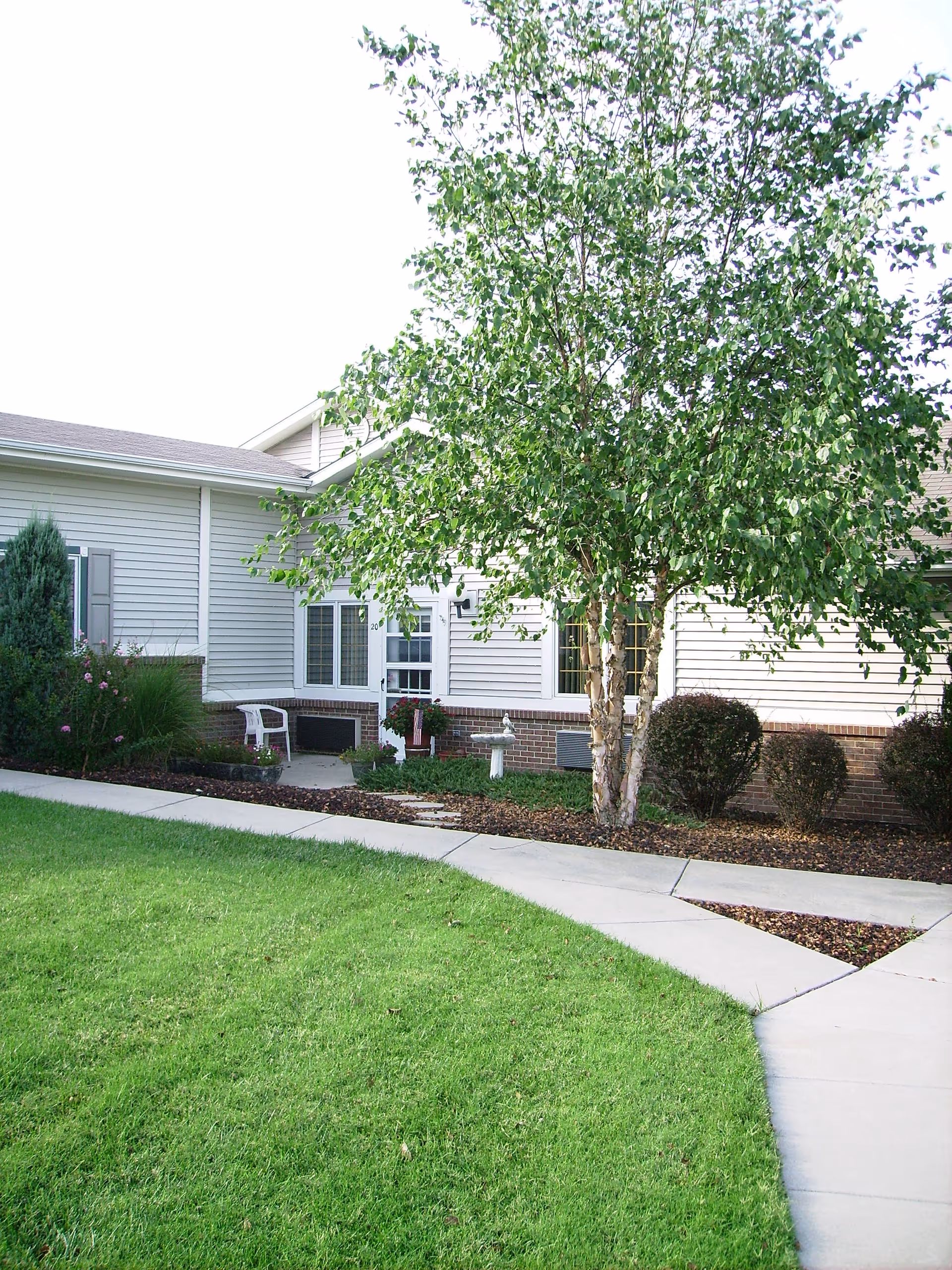 Front exterior of a single-story building with a paved walkway, green lawn, large tree, shrubs, and an entrance area with a chair.