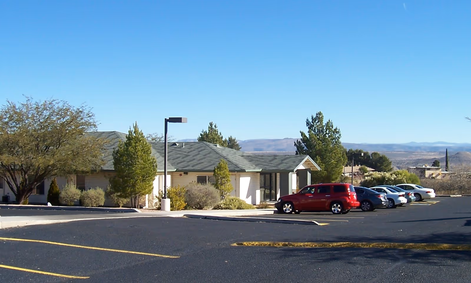 A single-story building with a green roof surrounded by trees and bushes, with a parking lot in front containing several parked cars under a clear blue sky.