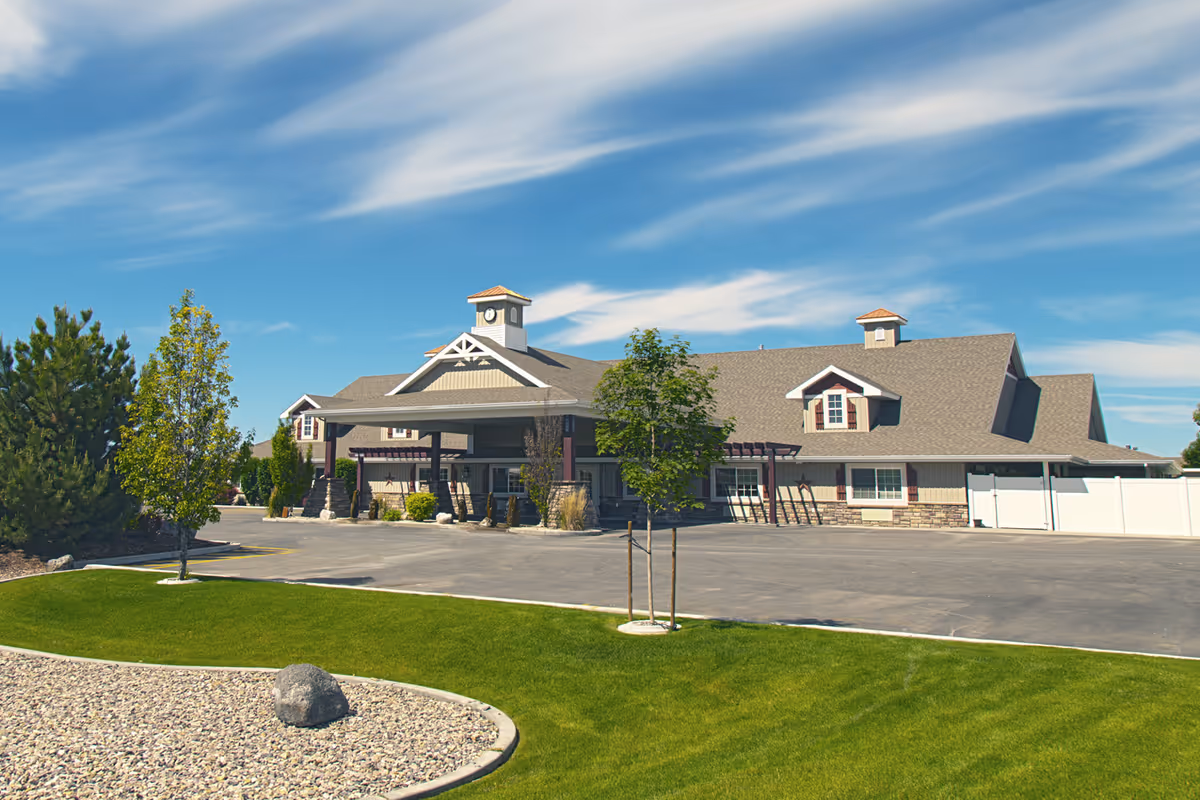 Exterior view of The Gables Memory Care of Blackfoot building under a blue sky with wispy clouds. The building has a gray roof with two cupolas, beige siding with stone accents, and a covered entrance. The foreground includes a paved driveway, green grass, small trees, and a landscaped area with rocks.