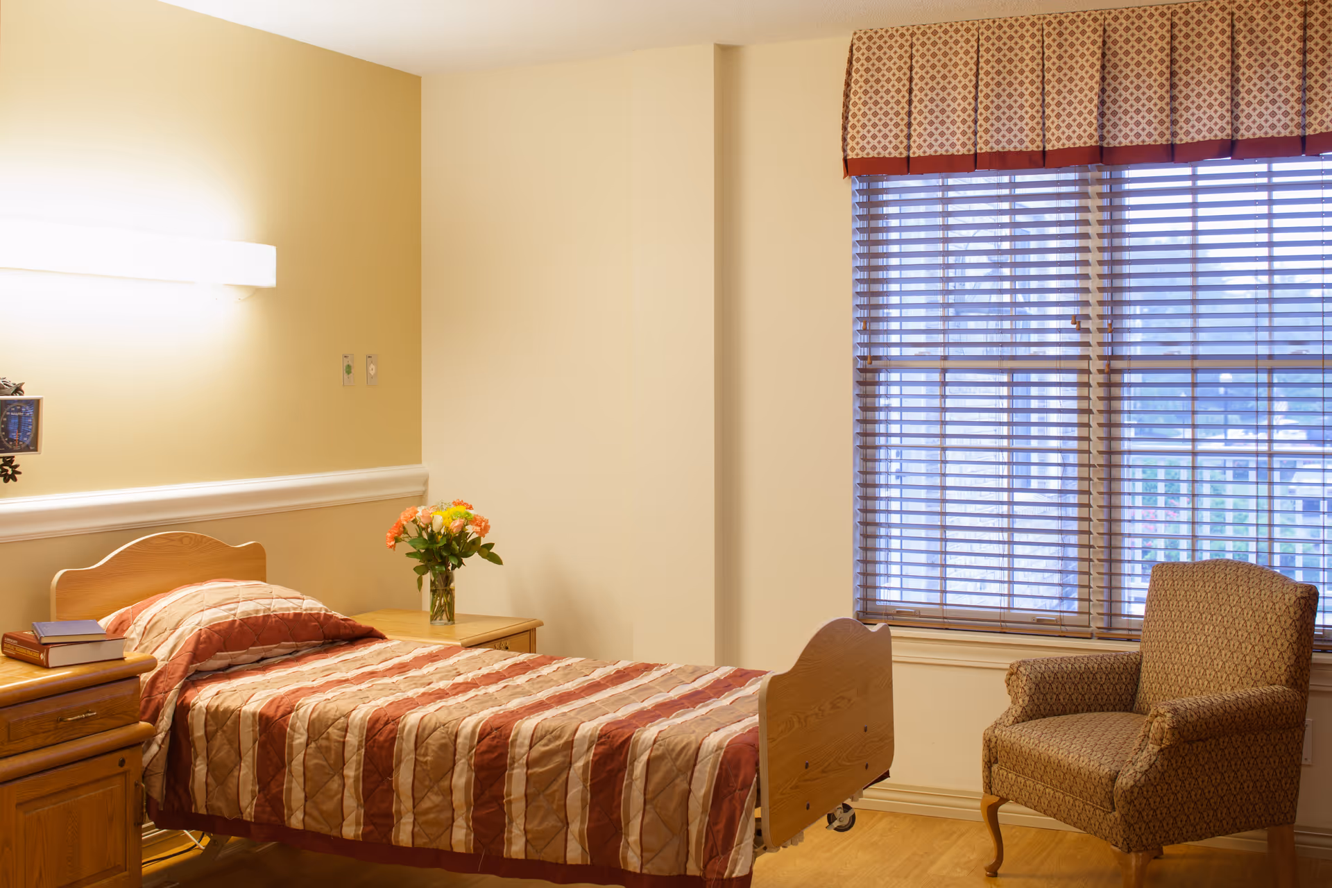 A single bed with a striped red and beige bedspread in a room with light yellow walls. There is a wooden nightstand next to the bed with a vase of flowers and some books. A patterned armchair is placed near a window with wooden blinds and a decorative valance.