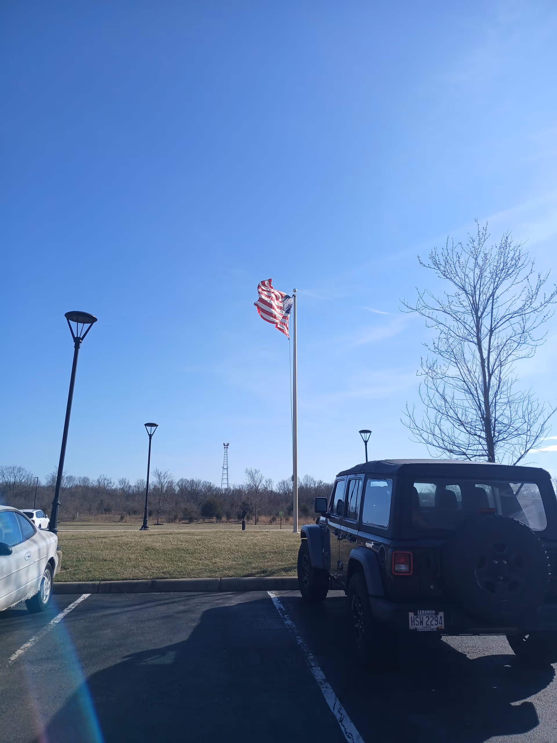 A parking lot with a Jeep, flagpole, American flag, leafless trees, and blue sky.