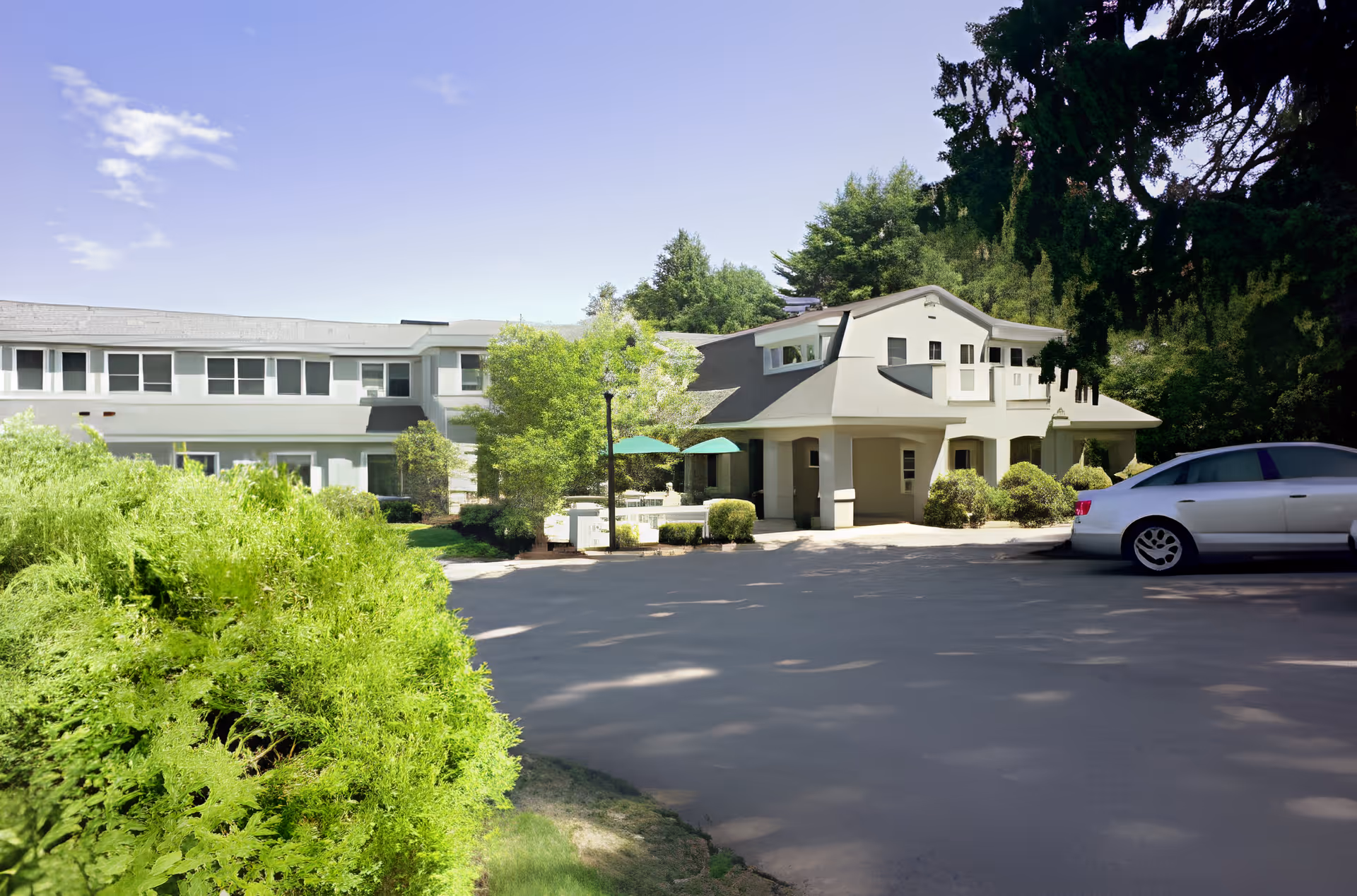 Front exterior of a light-colored senior living building with a driveway, parked car, and landscaped shrubs.