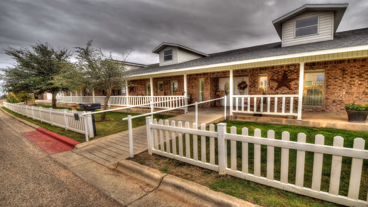 Brick senior living building with white fences, a ramp, and a covered front porch.