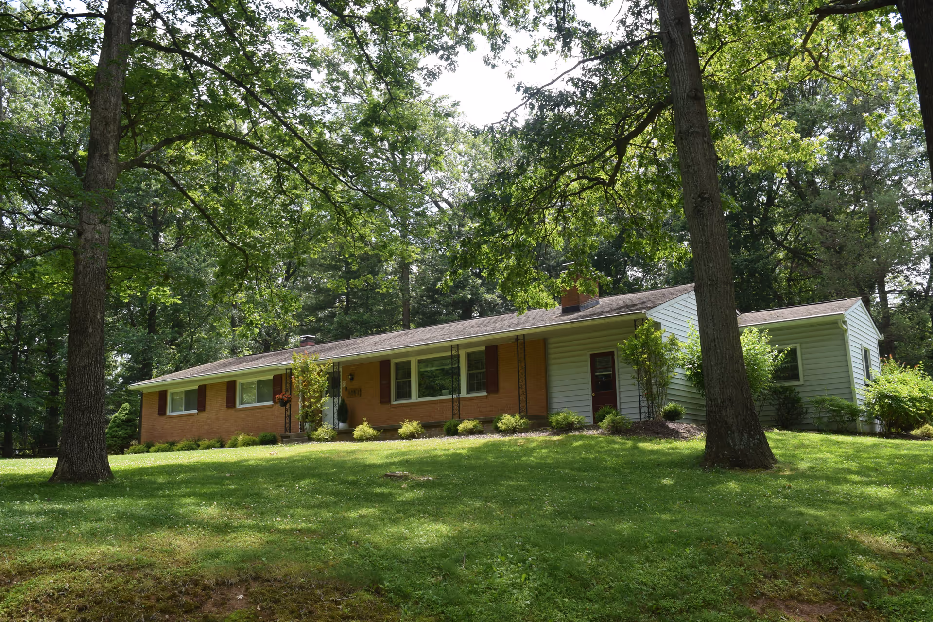 A single-story residential building with a brick and siding exterior surrounded by green grass and tall trees under a bright sky.