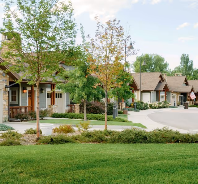 Row of senior living cottages with green lawns, trees, and shrubs along a curved street.