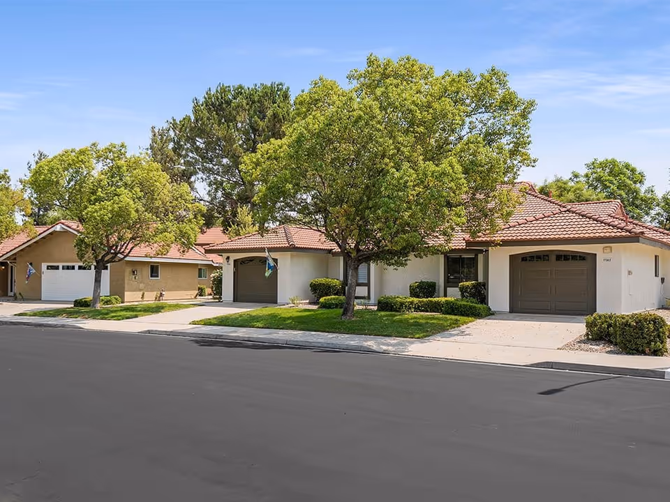 Single-story stucco homes with tile roofs, attached garages, and trees along a residential street.