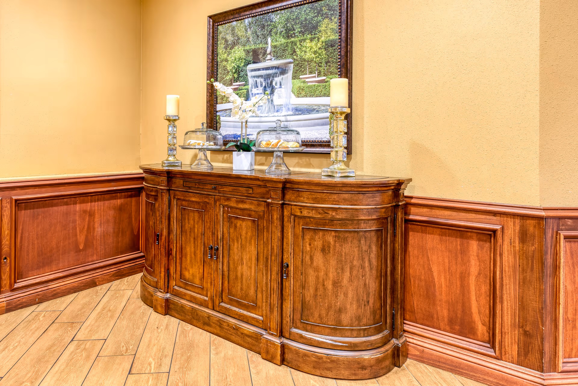 A wooden sideboard with a curved front and paneled doors sits against a beige wall with wooden wainscoting. On top of the sideboard are two glass cake stands with lids, a white vase with white flowers, and two tall candle holders with cream-colored candles. Above the sideboard hangs a framed picture of a fountain surrounded by greenery.