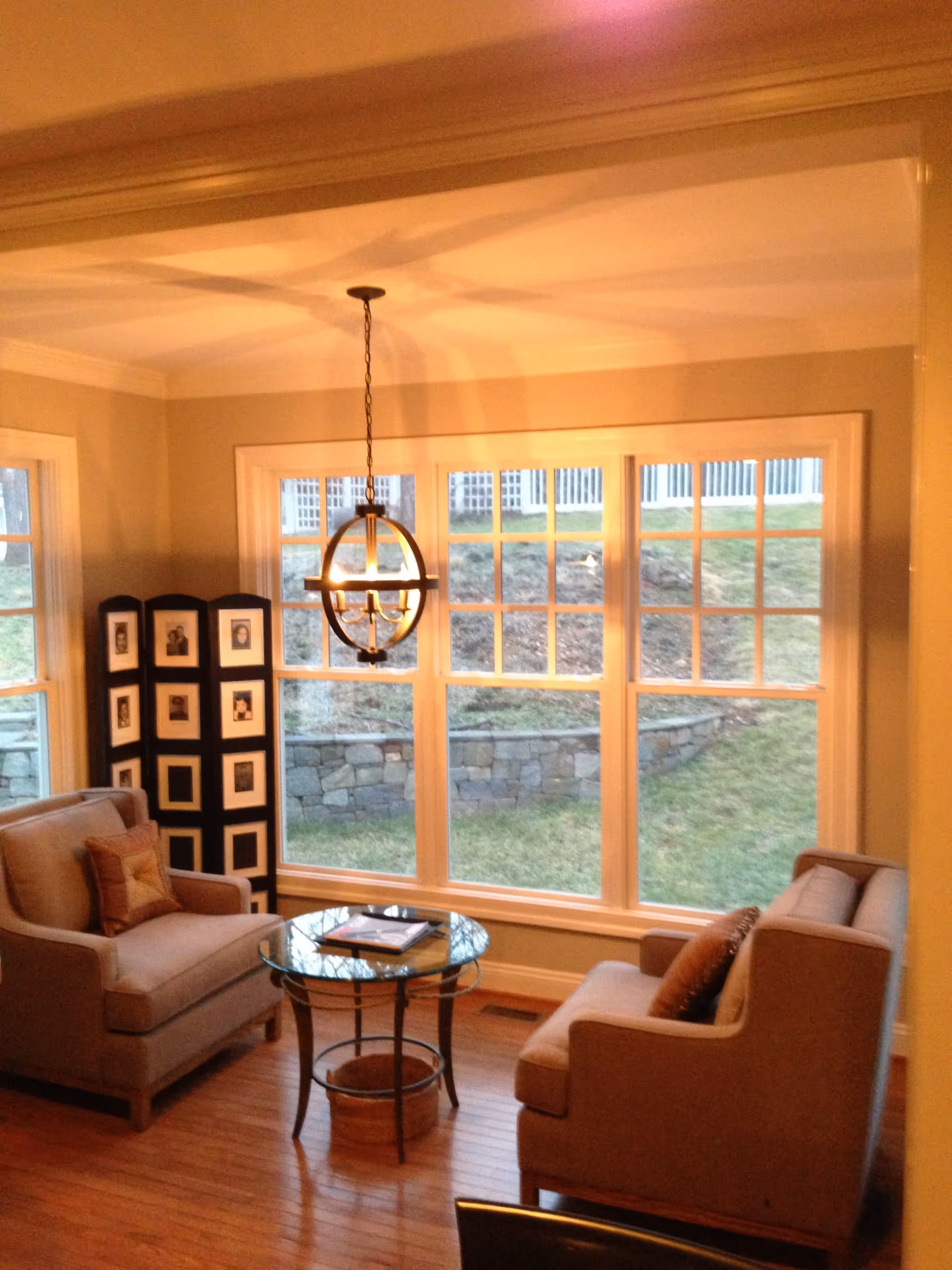 Cozy living room with two armchairs and a glass coffee table in front of large multi-pane windows under a hanging spherical chandelier.