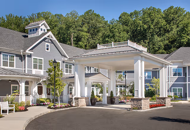 Front entrance of a gray senior living building with a covered porte-cochere, landscaped planters, and a paved driveway.