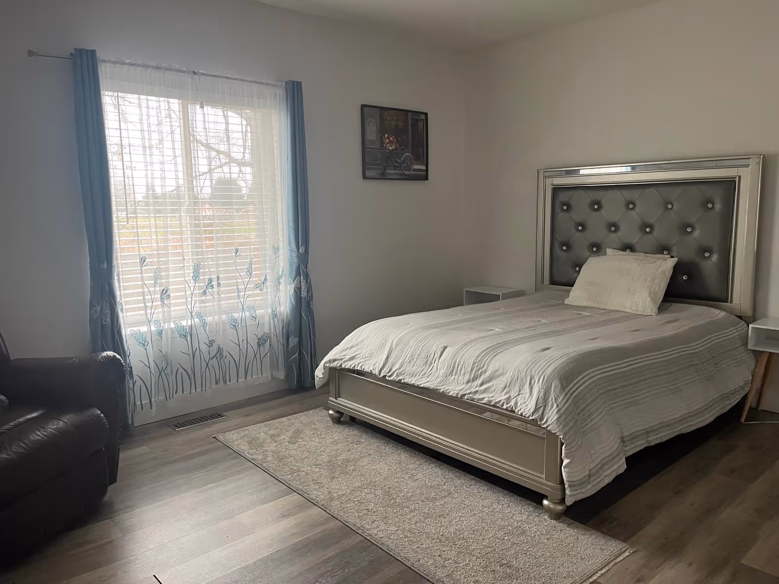A furnished senior living bedroom with a tufted headboard bed and blue-curtained window.