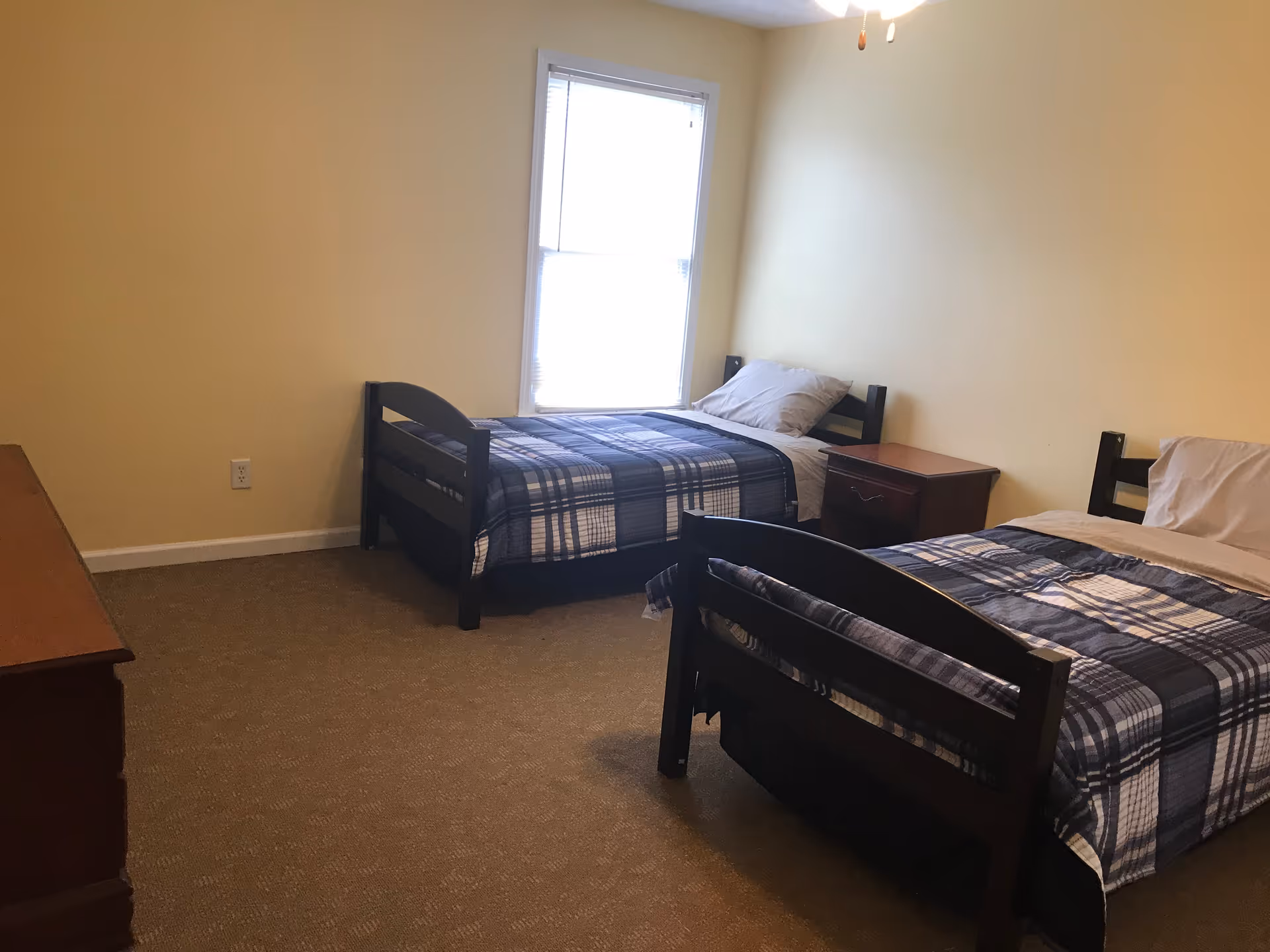 A simple bedroom with two single beds, each with a blue and white plaid blanket and a pillow. There is a wooden nightstand between the beds and a wooden dresser on the left side of the room. A window with blinds is centered on the far wall, allowing natural light into the room. The walls are painted light yellow and the floor is carpeted.