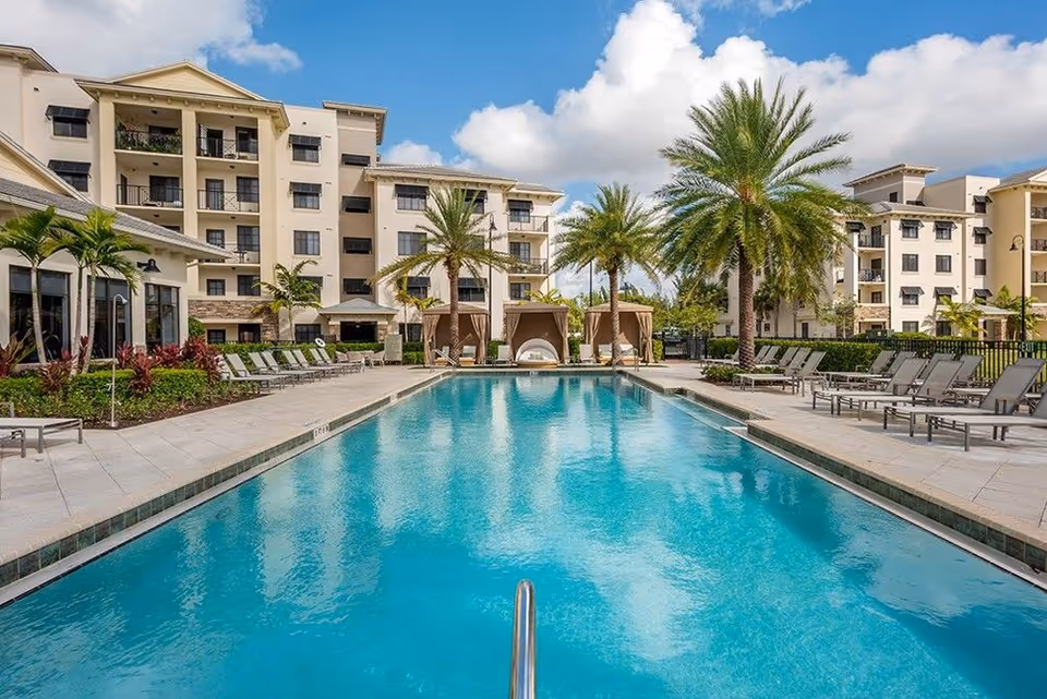 Outdoor swimming pool with lounge chairs, cabanas, palm trees, and multi-story residential buildings under a blue sky.