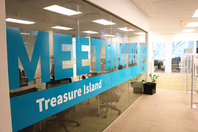 Glass-walled corridor with a large blue 'MEETING ROOM' sign and chair-filled offices.