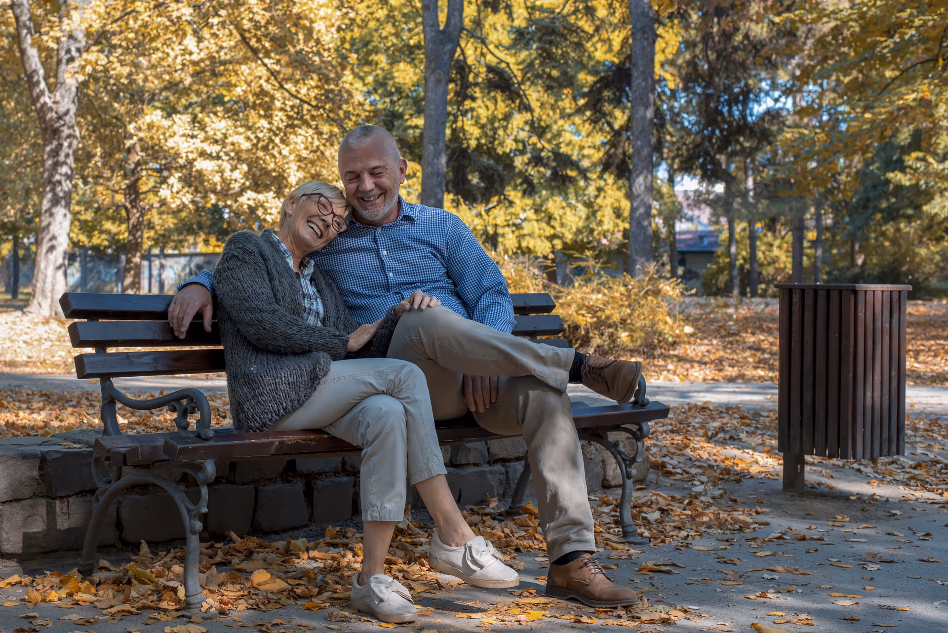 Couple sits on a park bench, smiling amid autumn leaves.