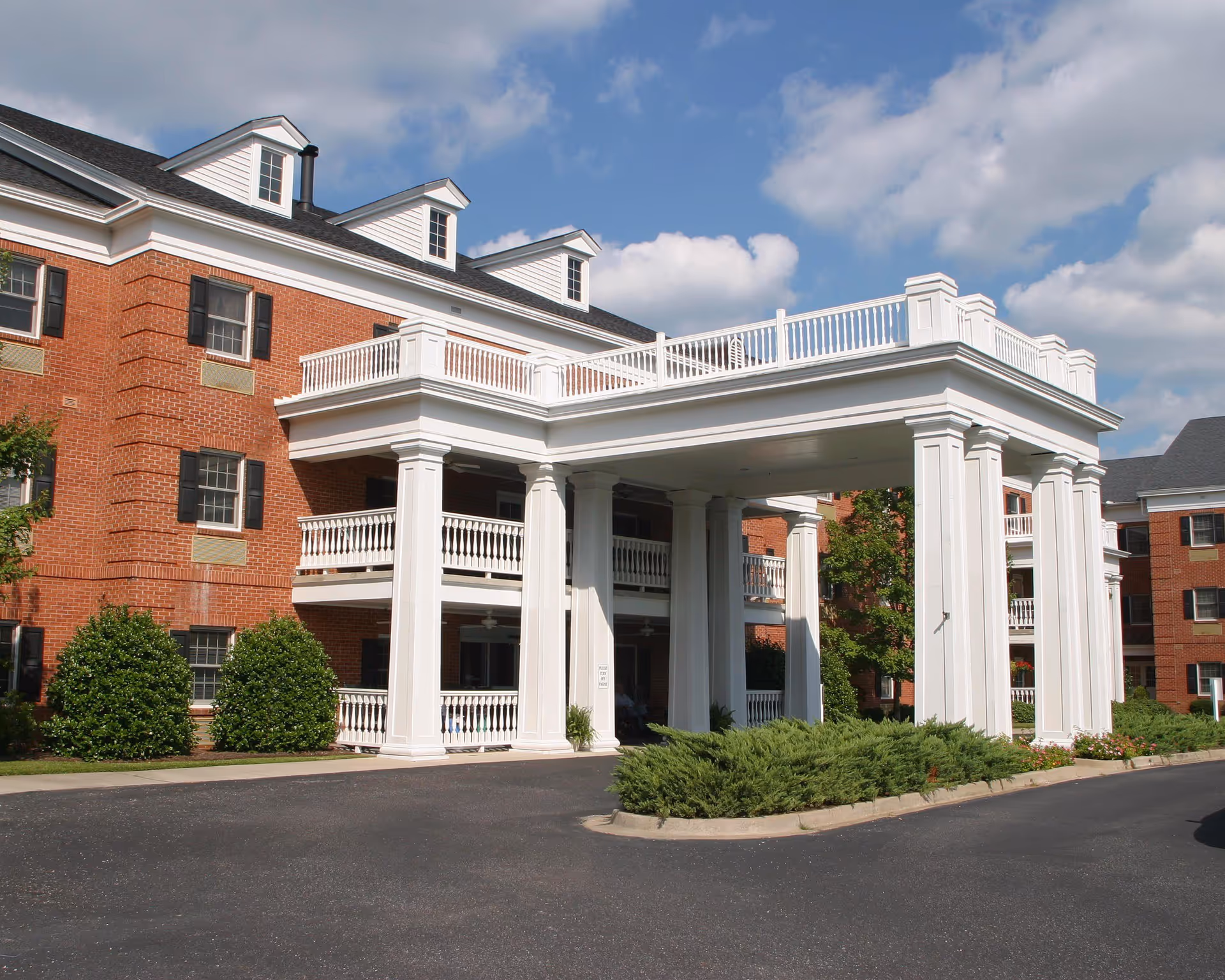 Exterior view of a senior living facility with red brick walls, white columns supporting a covered entrance, and balconies with white railings. The sky is partly cloudy and there are green shrubs and trees around the building.