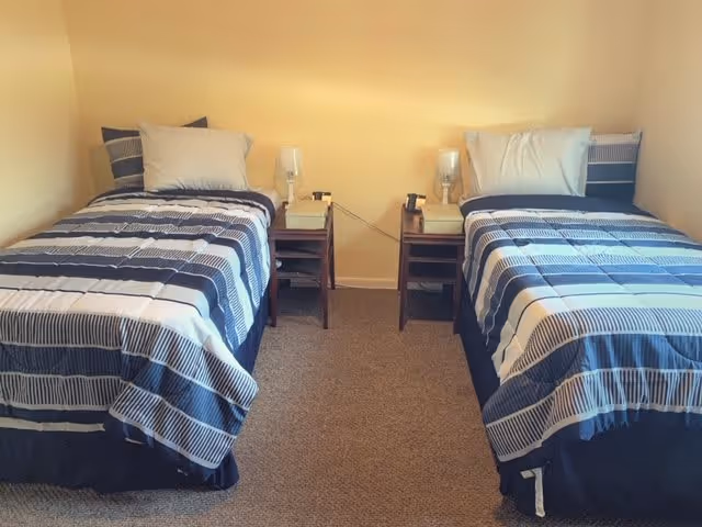 A bedroom with two twin beds placed parallel to each other against a beige wall. Each bed has a striped blue and white comforter and a white pillow. Between the beds are two small wooden nightstands, each with a lamp and a telephone on top. The floor is carpeted in a neutral color.