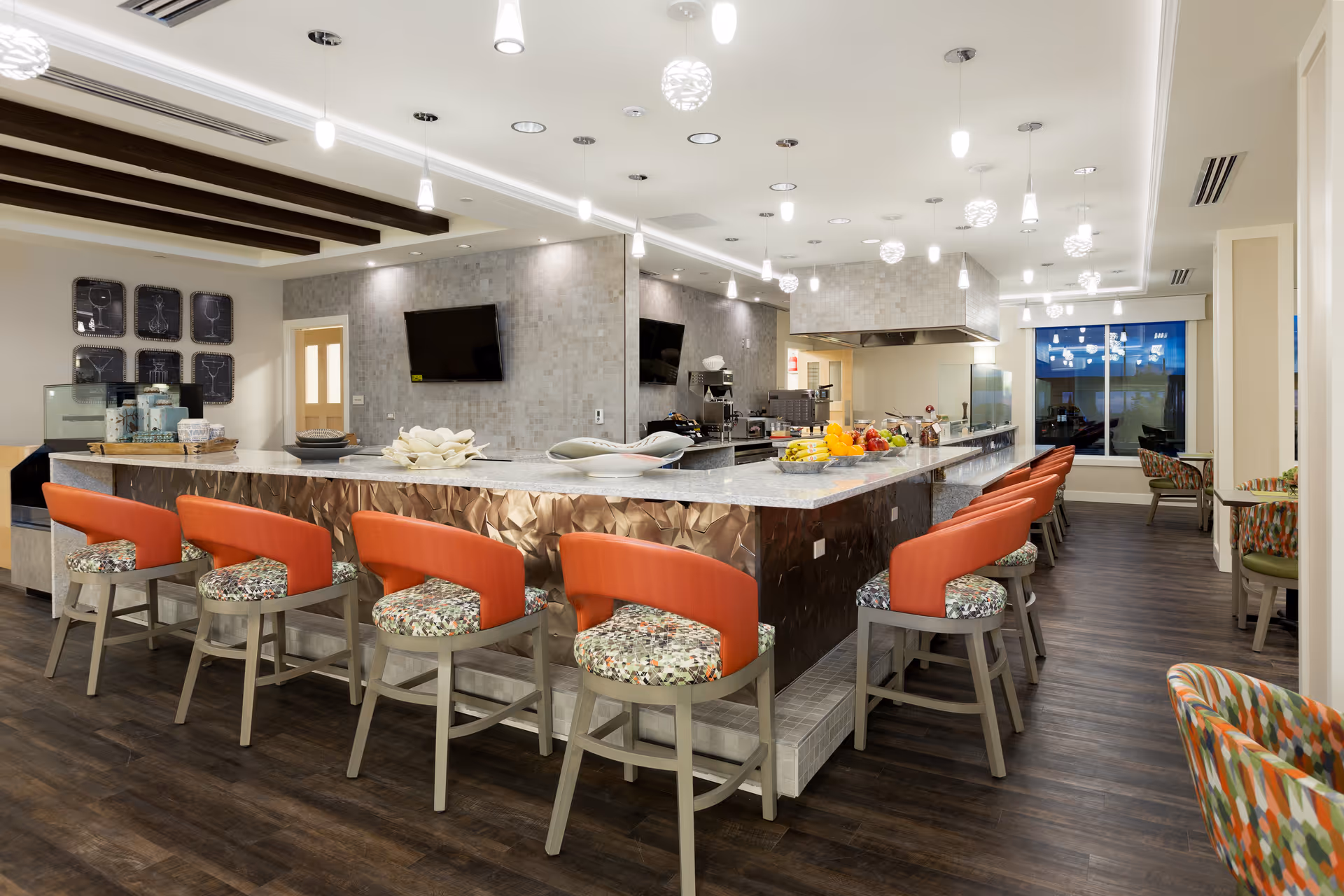 Modern kitchen and dining area with a large island counter surrounded by orange and patterned chairs. The kitchen features stainless steel appliances, a tiled backsplash, and pendant lighting. There are plates and fruit on the counter, and a television mounted on the wall in the background.
