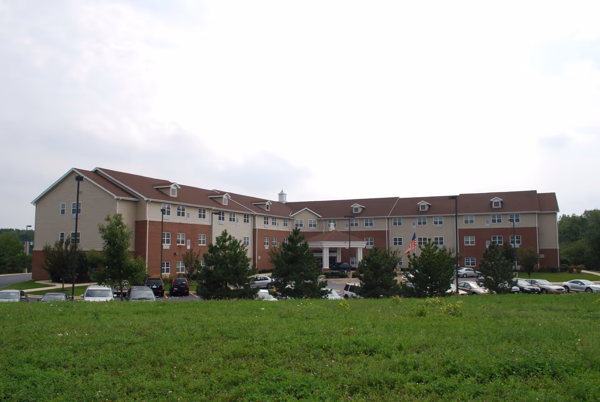 Exterior view of a large three-story senior living facility building with a combination of beige siding and red brick. The building has multiple windows and a covered entrance with white columns. There are several cars parked in front, an American flag on a flagpole, and green grass and trees in the foreground under a cloudy sky.