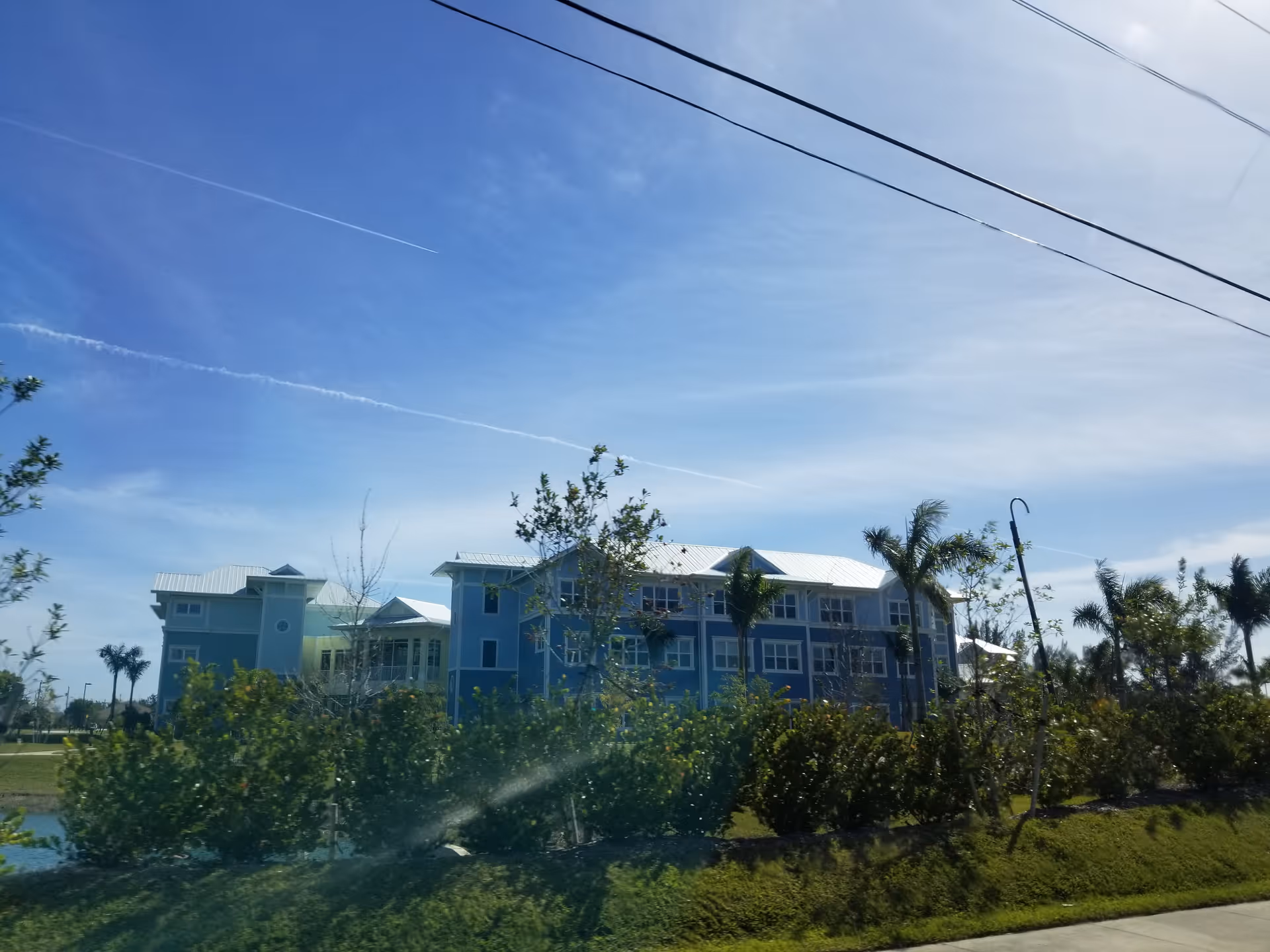 Blue multi-story senior living building with white roof, palm trees, landscaping, and power lines overhead