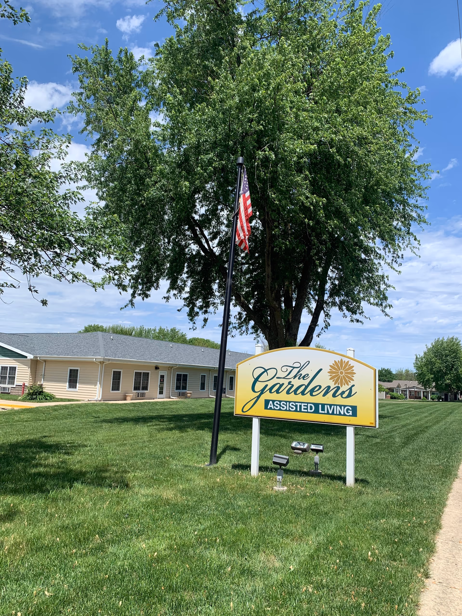 A large green lawn with a sign that reads 'The Gardens Assisted Living' in front of a single-story beige building with a gray roof. There is a tall tree behind the sign and an American flag on a flagpole next to it. The sky is partly cloudy with blue patches.