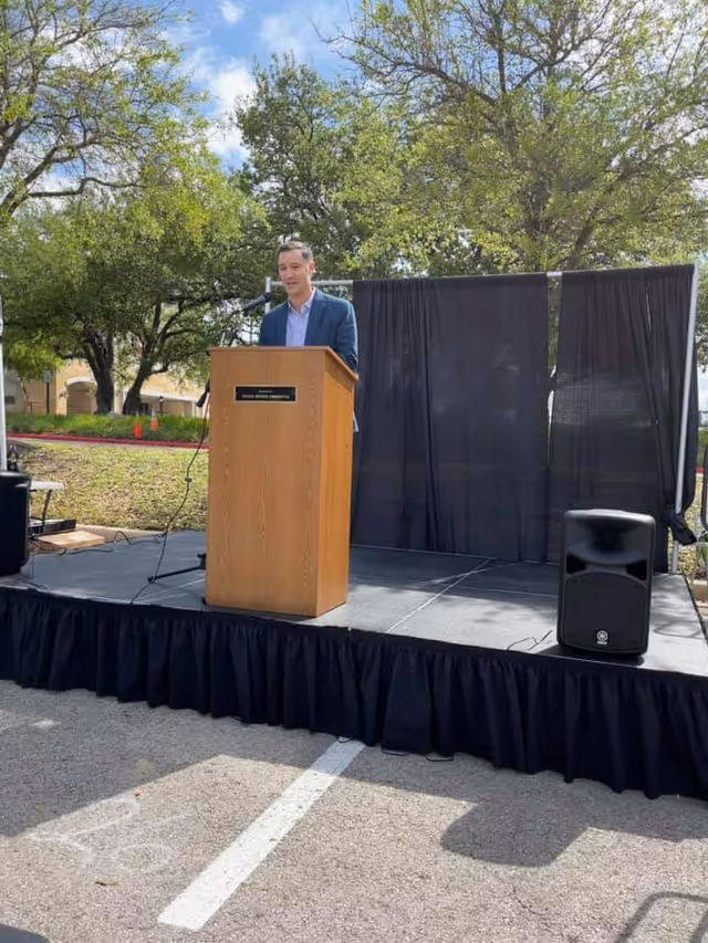 Man in a blue suit speaks at a wooden podium on an outdoor stage.