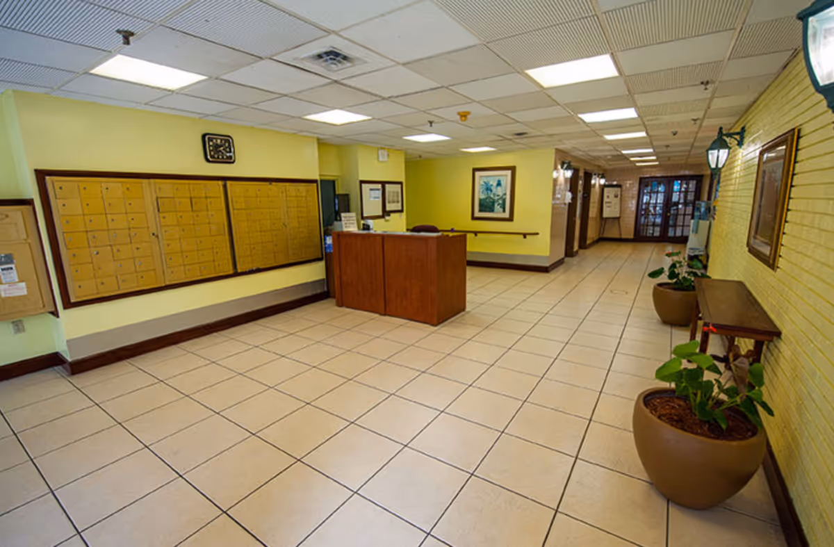Reception lobby with a central wooden desk, wall mailboxes, tiled floor, and potted plants.