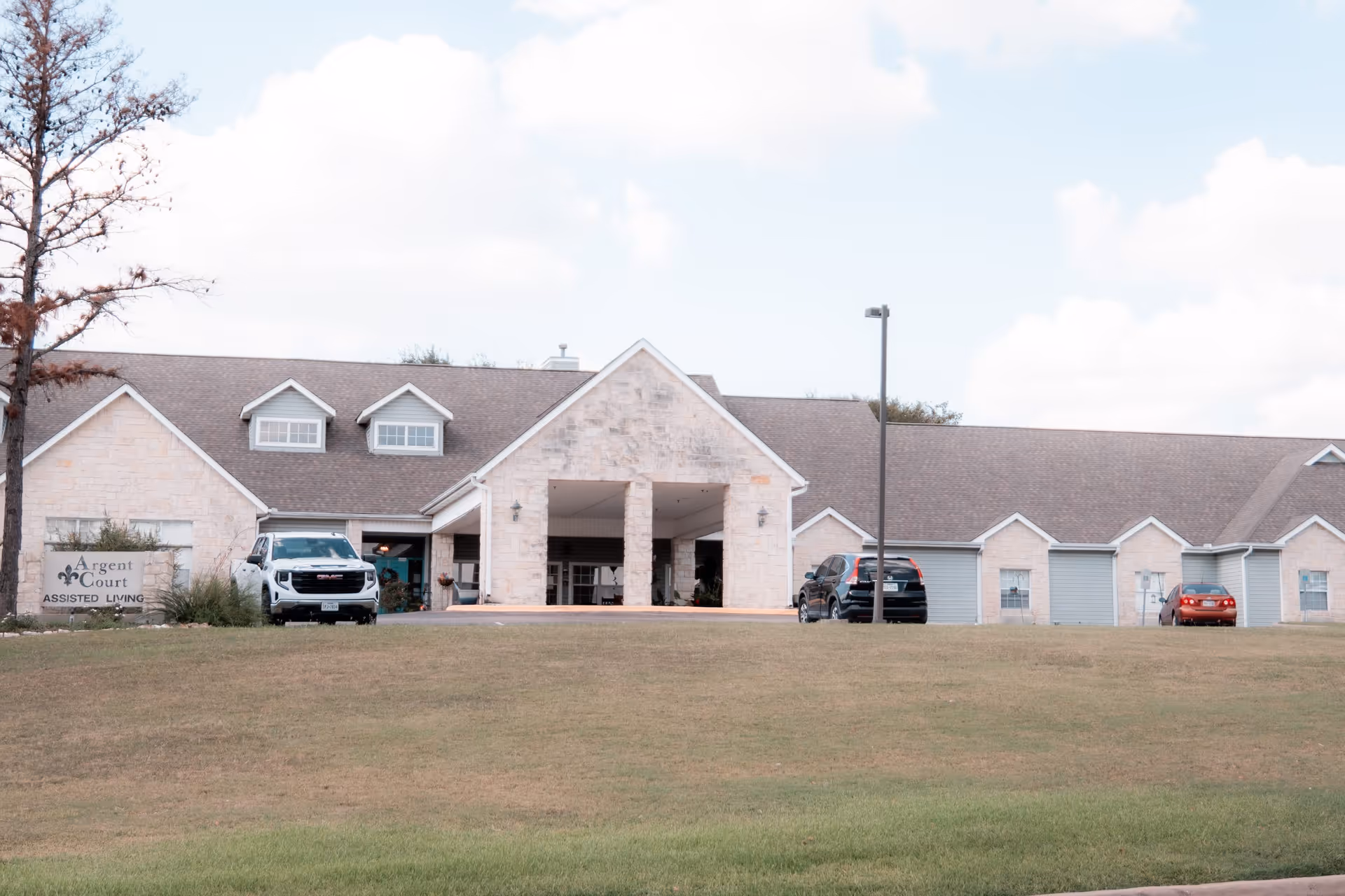Front exterior of Argent Court Assisted Living showing the building entrance, parked cars, and a grassy lawn.
