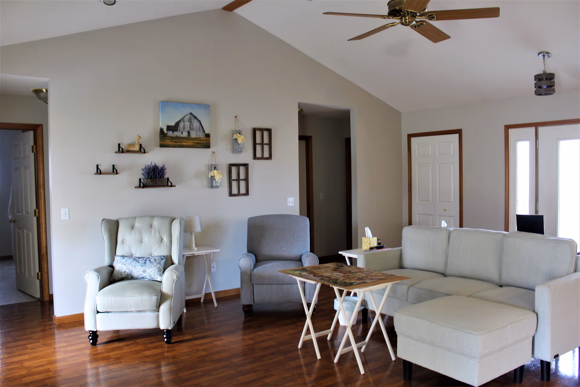 A living room with a light gray sectional sofa with an ottoman, a gray armchair, and a light green tufted armchair with a decorative pillow. There are two small white side tables, one with a lamp and the other with a tissue box. A small folding table with a puzzle on it is in front of the seating. The walls are light gray with rustic wall decor including a painting of a barn, small shelves, and hanging flower jars. The floor is wood, and there is a ceiling fan with wooden blades above.