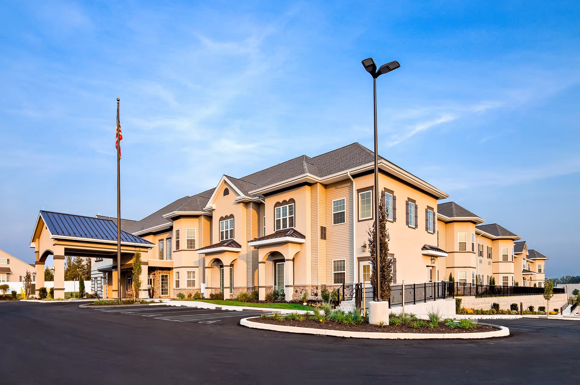 Two-story beige senior living facility with a covered entrance, flagpole, and landscaped parking area under a blue sky.