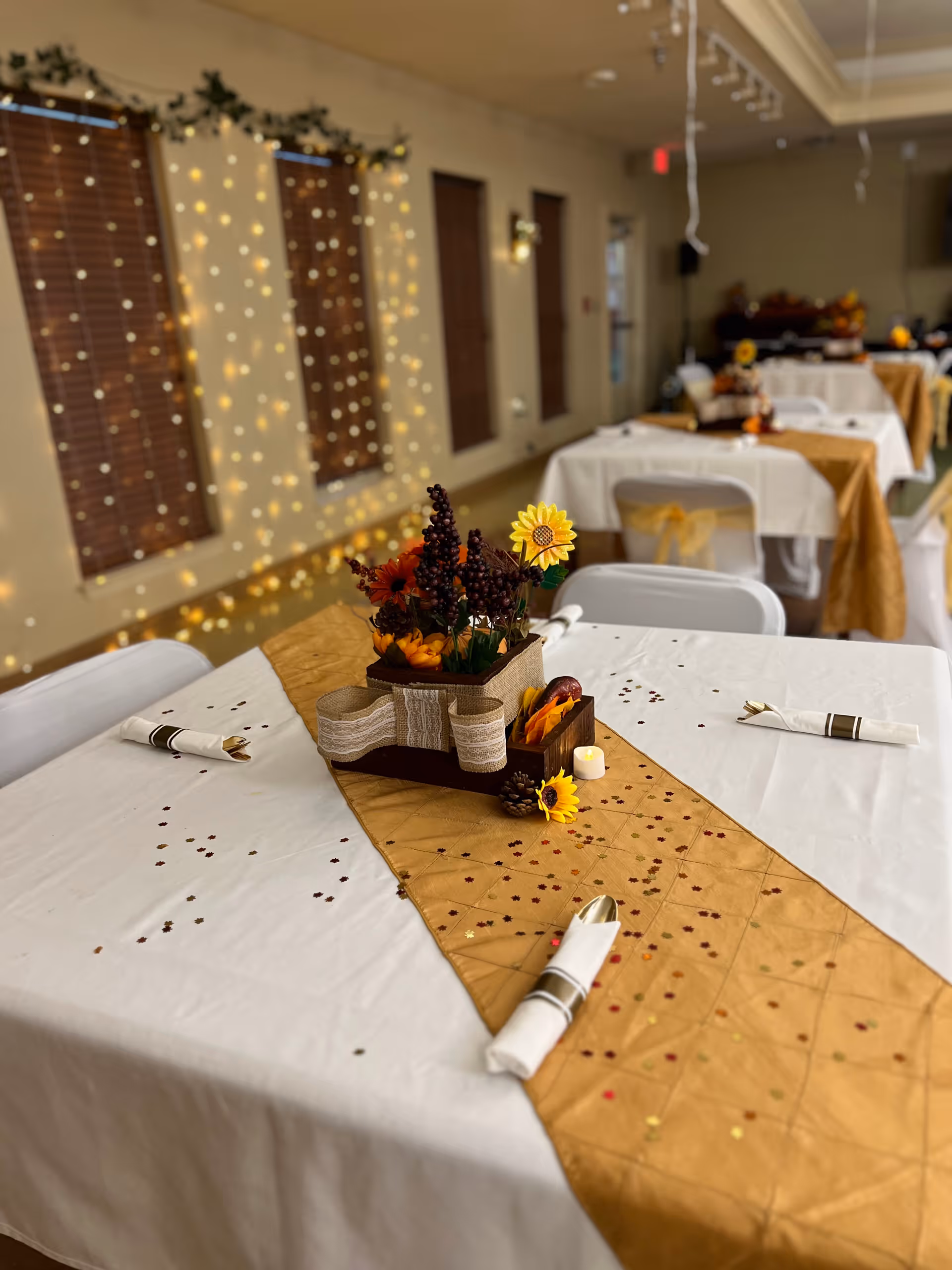 Decorated dining room tables with white tablecloths, gold runners and autumn floral centerpieces set for an event with string lights on the wall.
